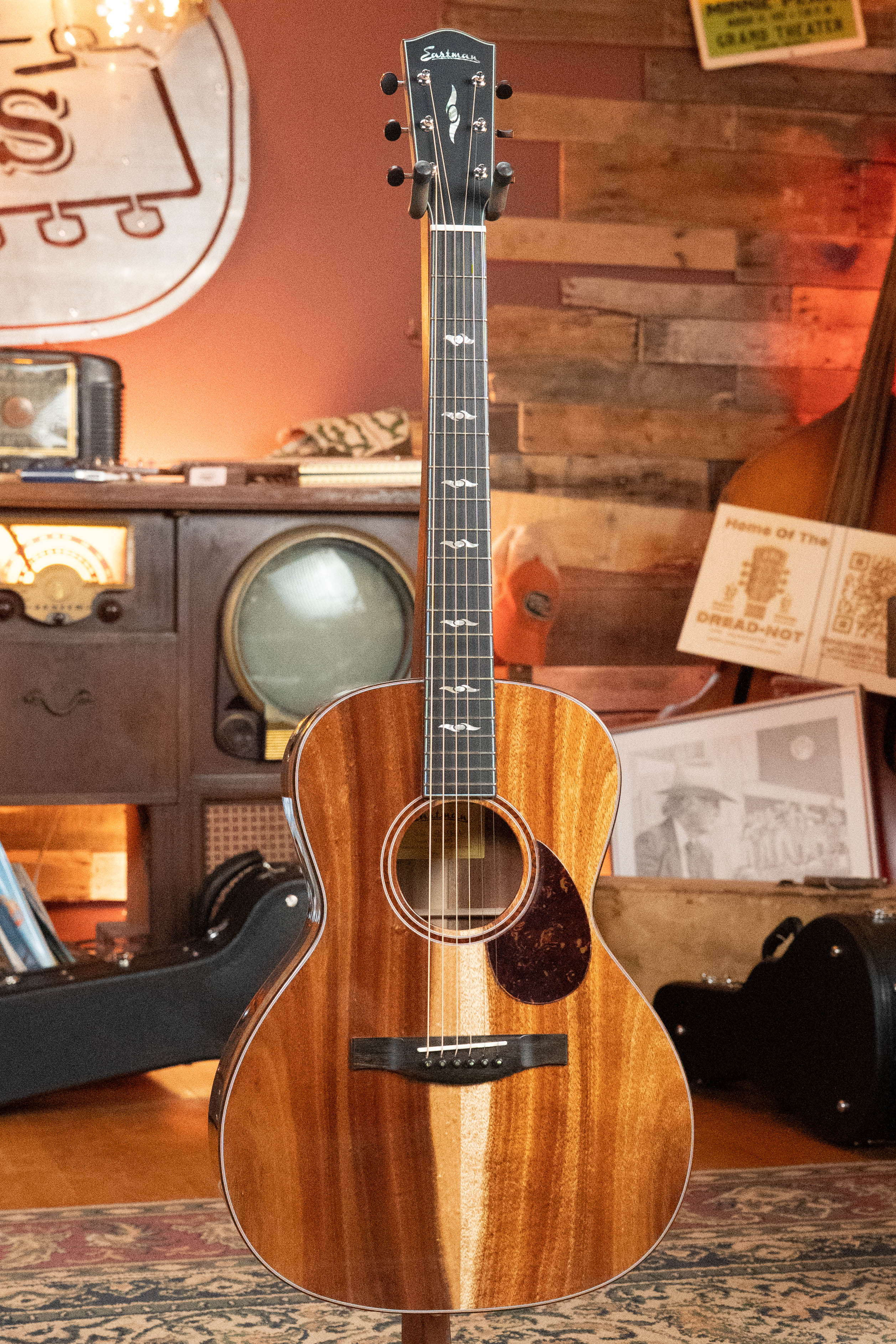 A (Used) 2025 Eastman Guitars L-OOSS-LTD-KOA Limited Edition All Koa Acoustic Guitar #9075 with a glossy finish stands upright in a cozy, vintage-themed room featuring a retro TV, records, and wood-paneled walls.