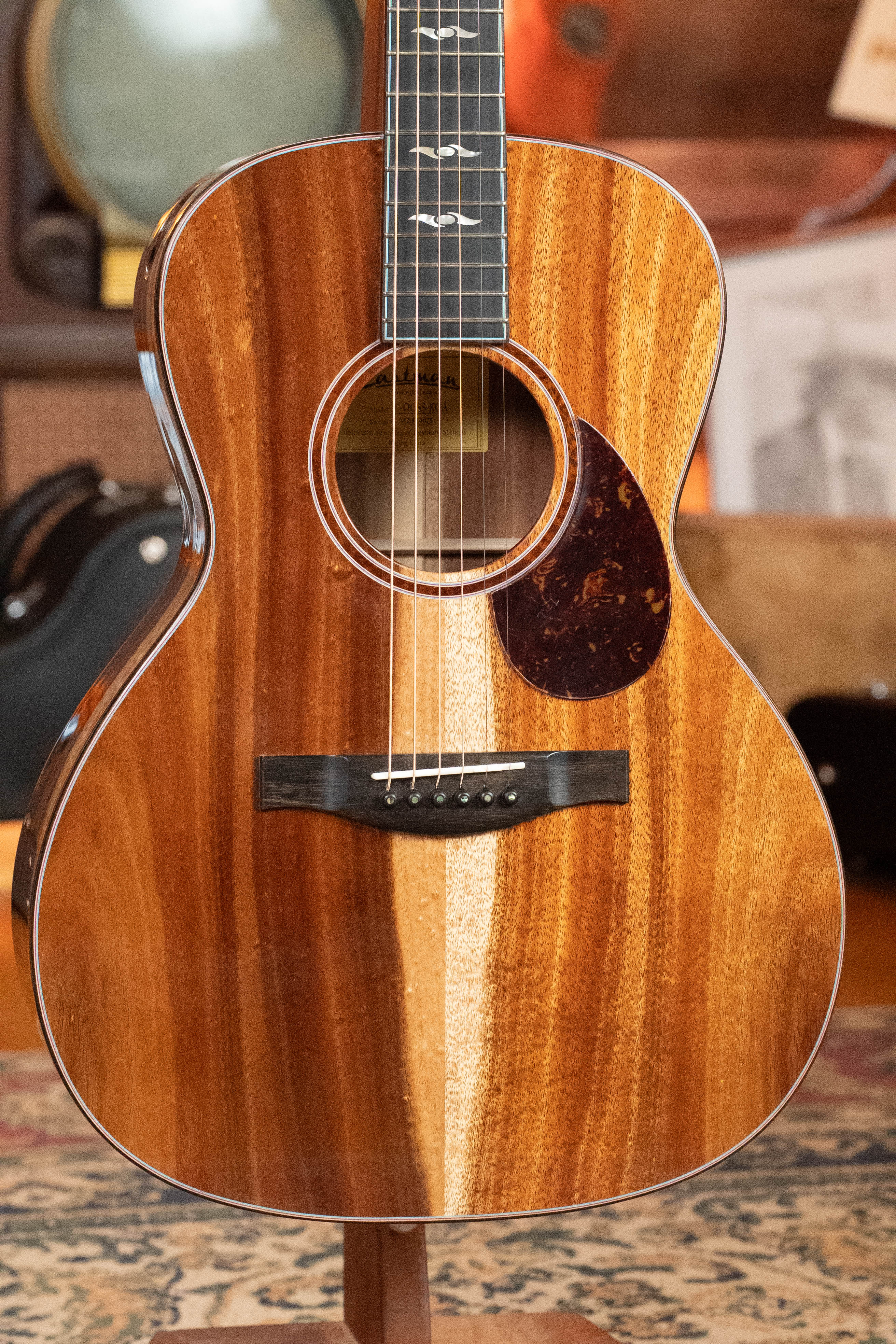 A close-up of a used 2025 Eastman Guitars L-OOSS-LTD-KOA Limited Edition Luthier Series all-koa acoustic guitar (#9075) with a glossy finish, dark tortoiseshell pickguard, subtle bird fretboard inlays, displayed indoors on a stand.