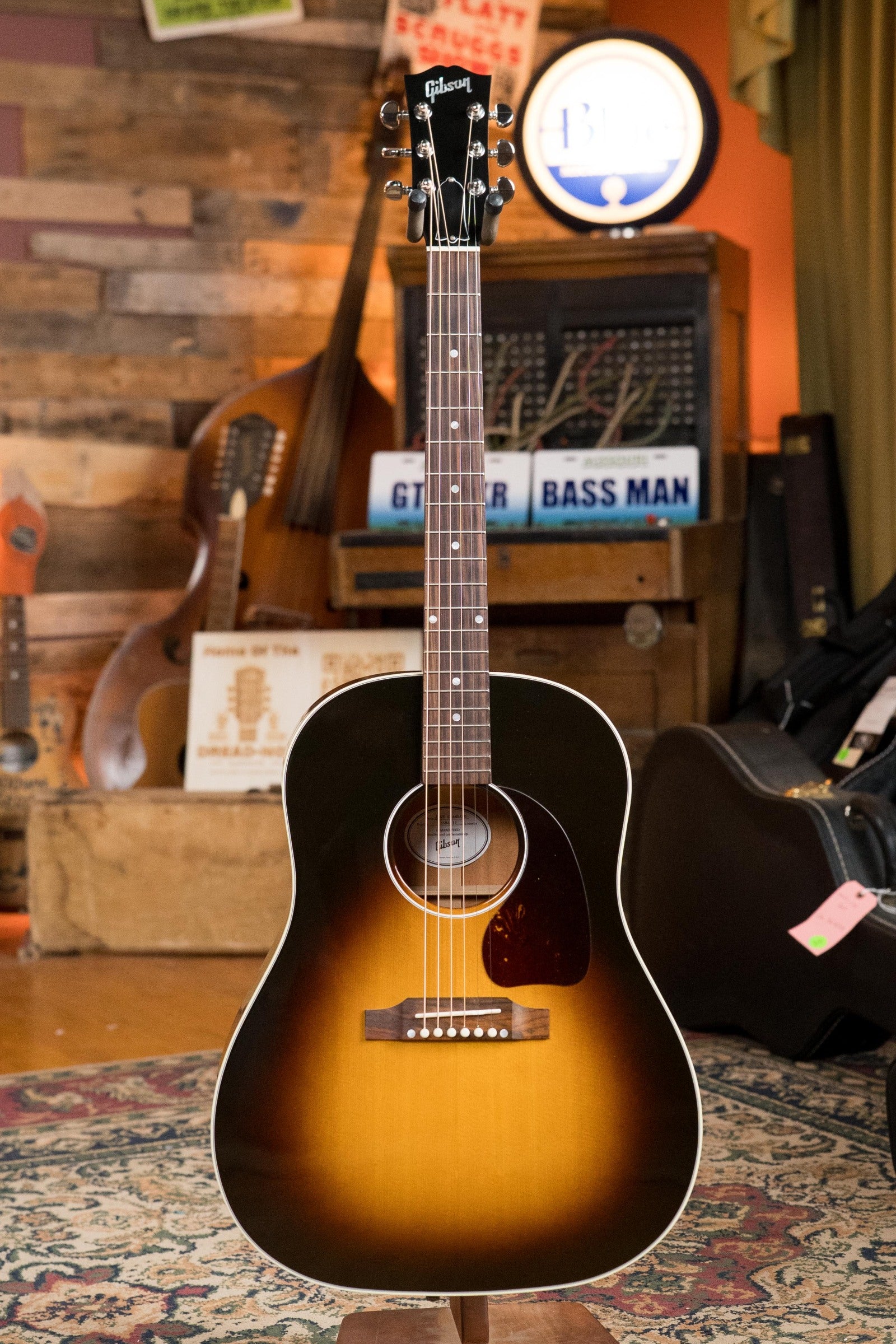 A used Gibson J45 Standard Sitka/Mahogany Vintage Sunburst Dreadnought with K&K Pickup (#4111) stands upright in a cozy room with wooden walls, vintage gear, and instrument cases in the background.