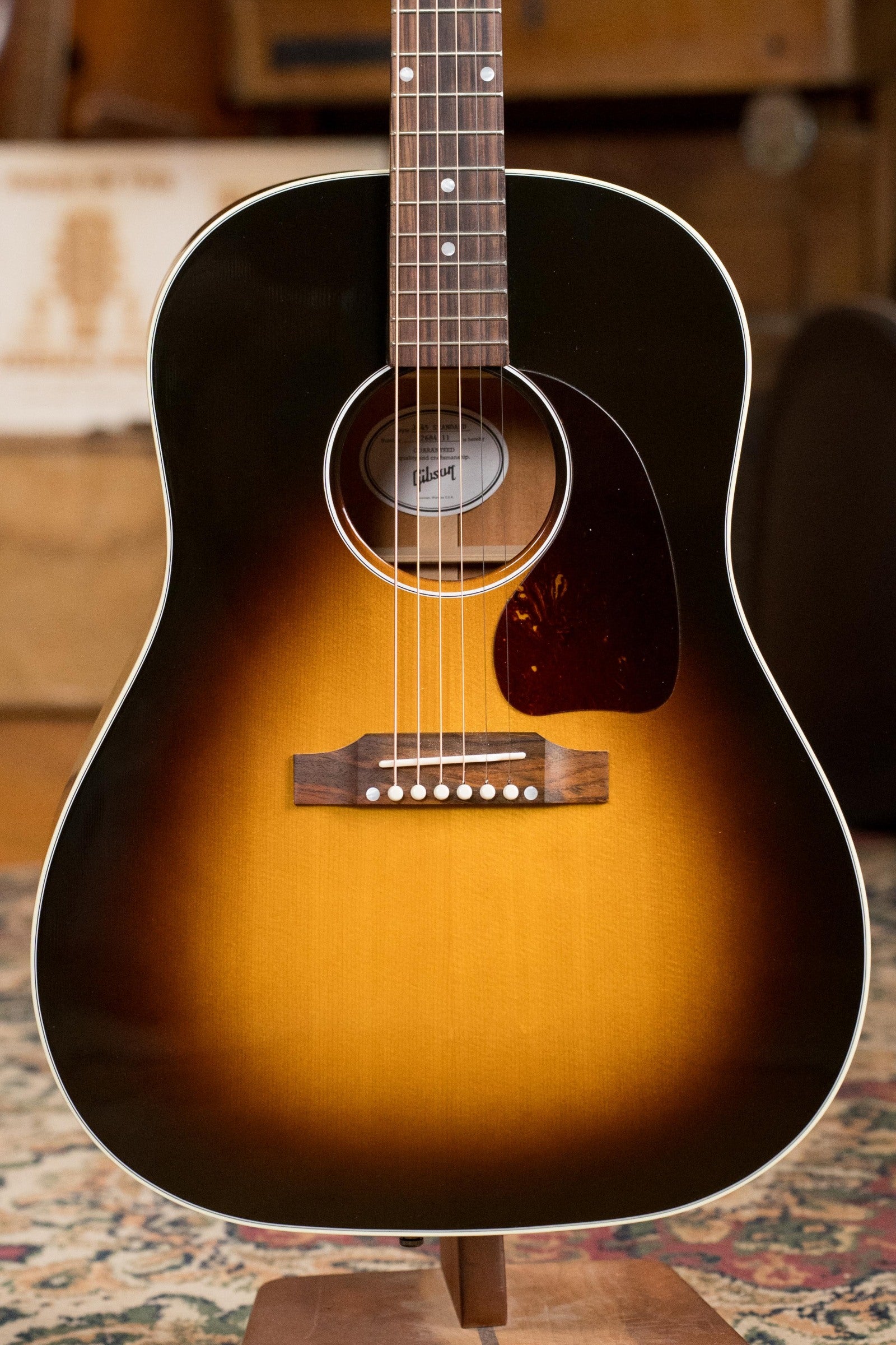 A used Gibson J45 Standard Sitka/Mahogany Round Shoulder Vintage Sunburst dreadnought guitar with K&K pickup (#4111) stands upright on a patterned rug.