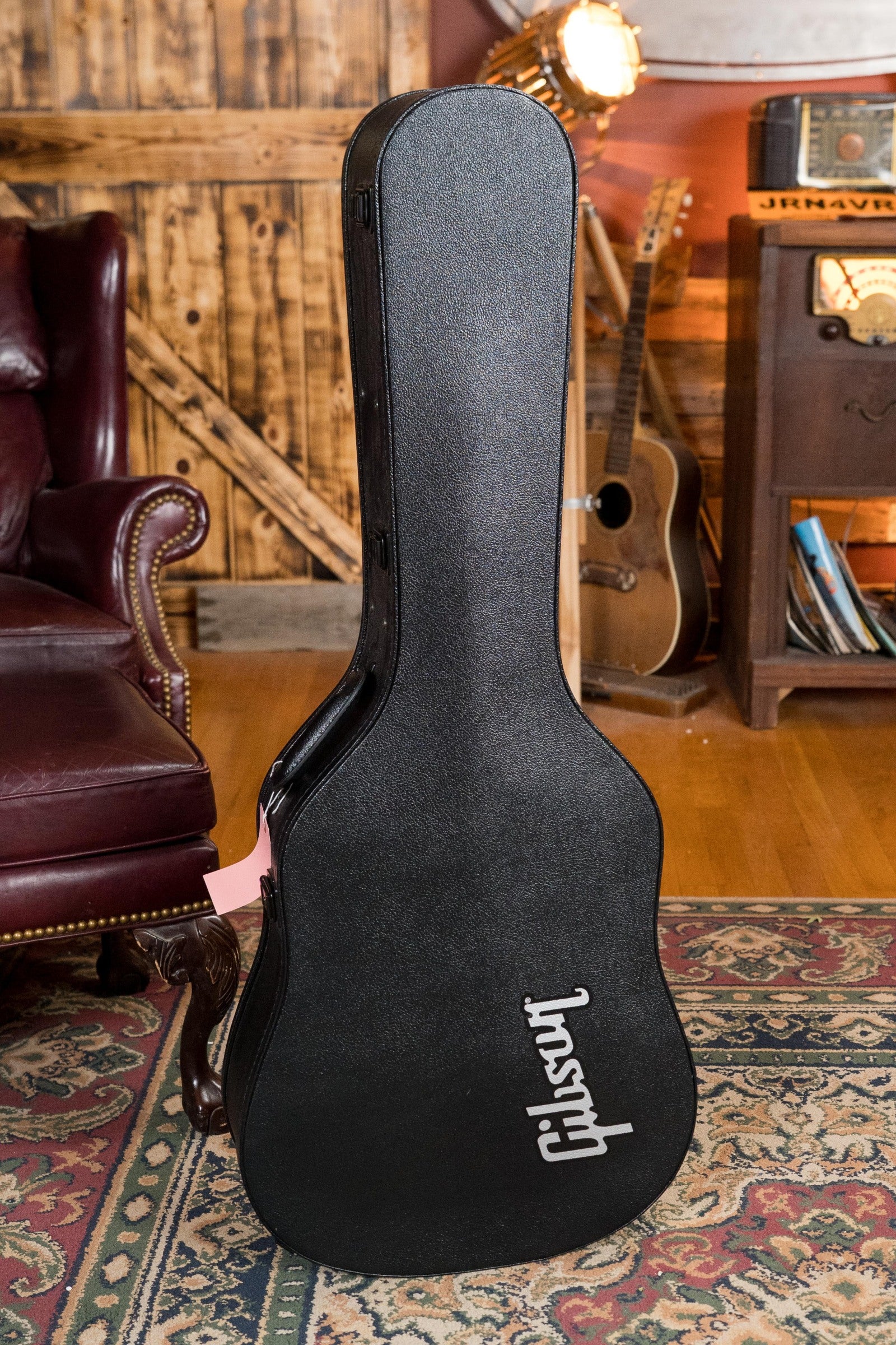 A black Gibson case stands upright on a rug in a cozy room, with leather chair and vintage furniture—ideal for showcasing your used Gibson J45 Standard Sitka/Mahogany Vintage Sunburst Dreadnought (#4111) w/ K&K Pickup.