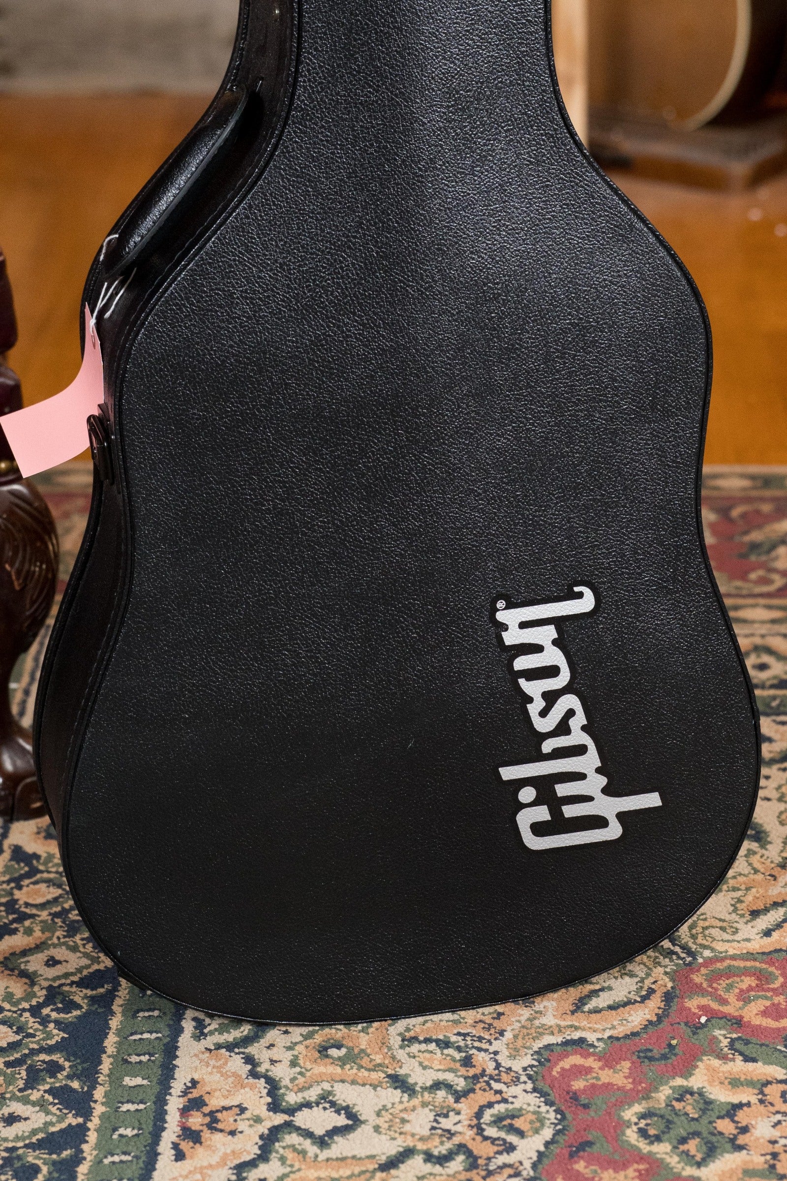 A black Gibson guitar case with a white logo rests on a patterned rug, holding a used Gibson J45 Standard Vintage Sunburst Dreadnought (#4111) with K&K pickup; a pink tag hangs from the handle and some wooden furniture is partly visible behind.