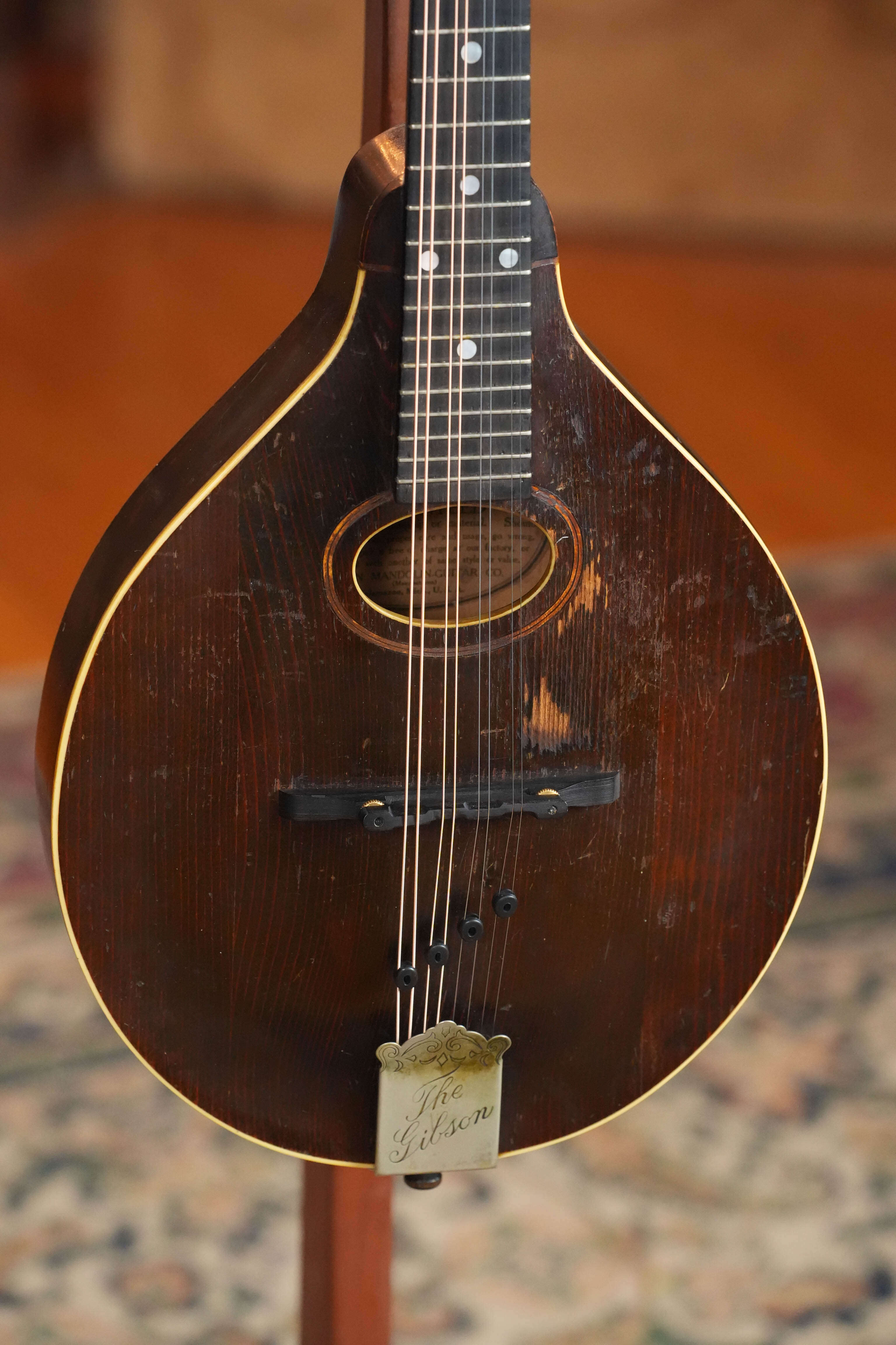 A close-up of a (Used) Vintage 1920 Gibson Spruce/Birch Oval-Hole Teardrop A-Style Mandolin #9066 with dark wood finish and engraved metal tailpiece, resting on a patterned rug.