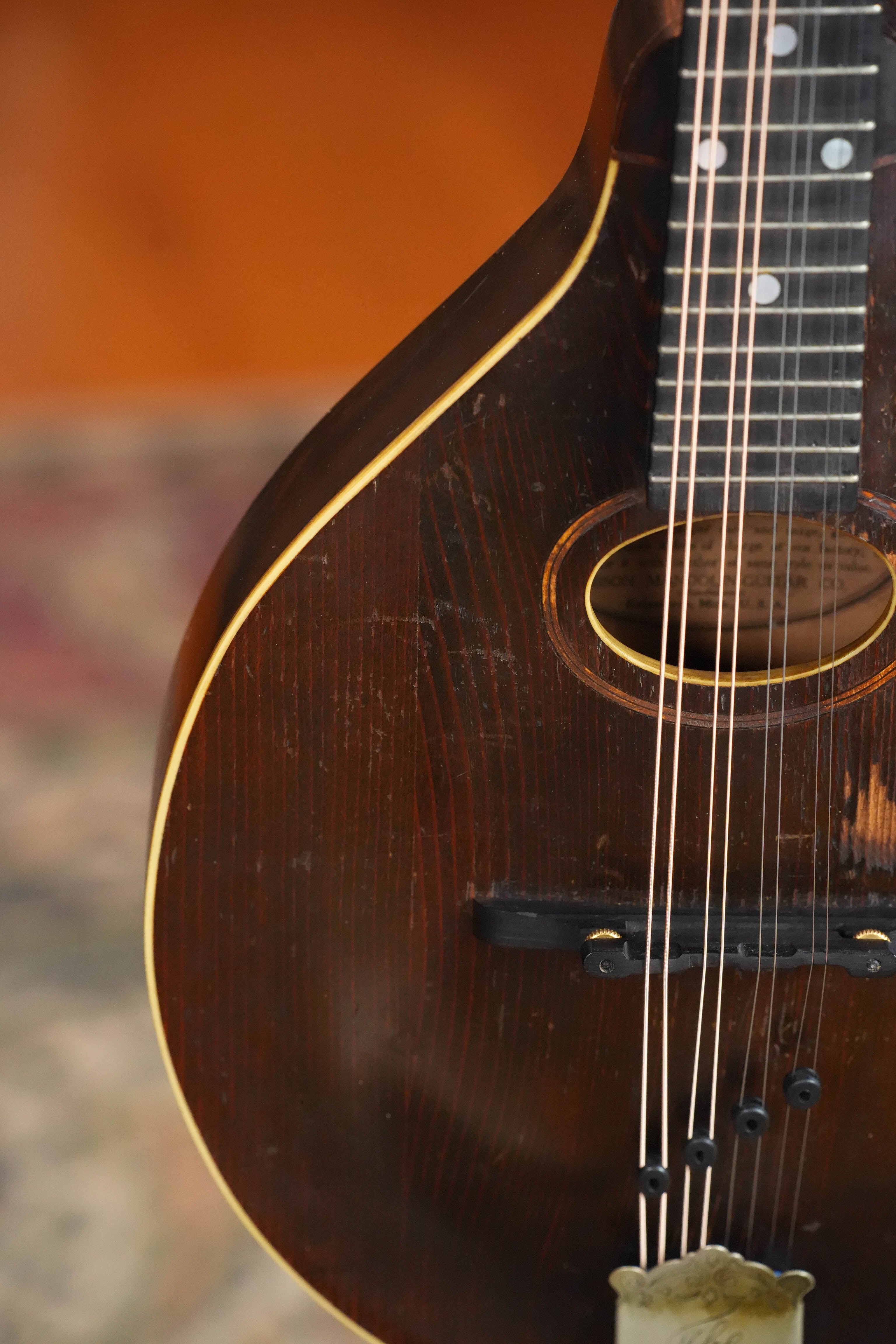 A close-up of a (Used) Vintage 1920 Gibson Spruce/Birch Oval-Hole Teardrop A-Style Mandolin #9066 shows its strings, sound hole, and neck against a blurred rug and warm-toned wall.