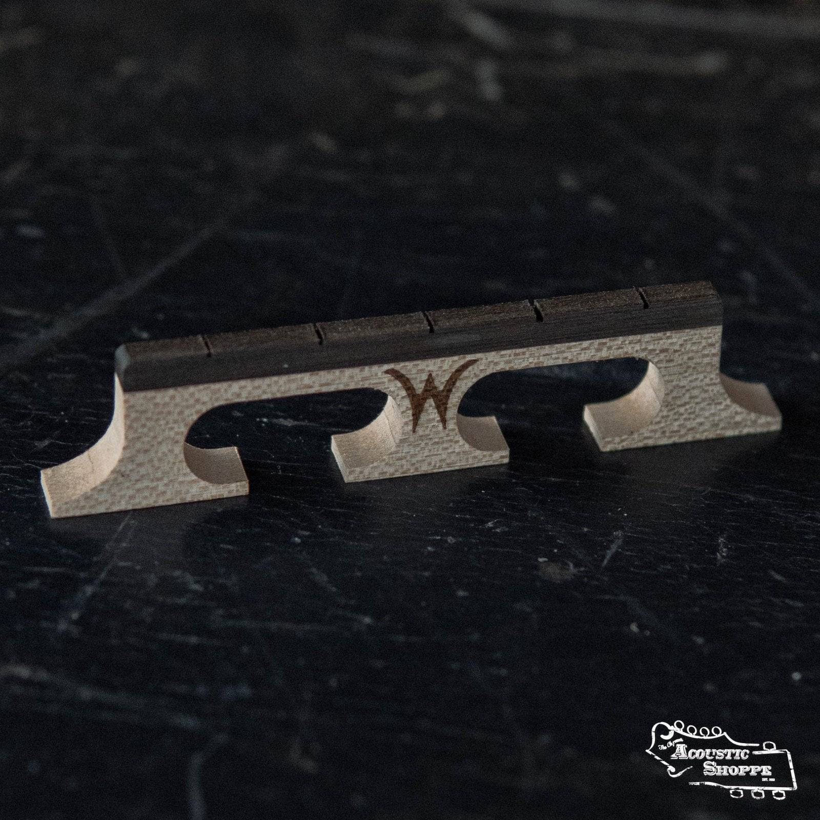 The Wyatt Custom Banjos Red Maple 5/8 Banjo Bridge with Crowe spacing features three arches, a dark top strip, and a central stylized W logo. It rests on a dark surface with The Acoustic Shoppe logo at the bottom right.