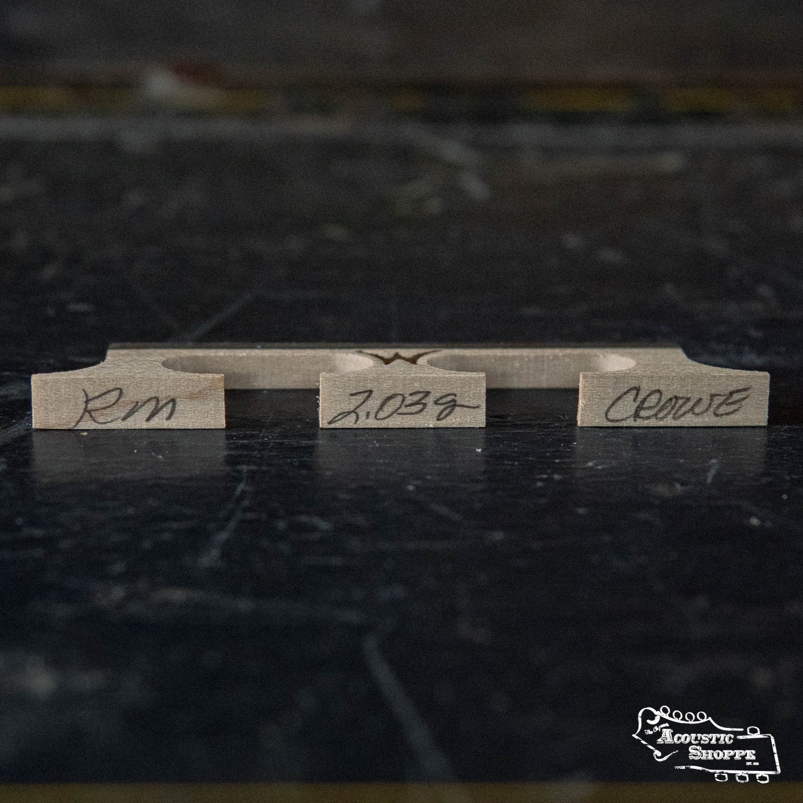 A Wyatt Custom Banjos Red Maple 5/8 Banjo Bridge with Crowe spacing and handwritten markings (RM, 2.039, Clewe) sits on a dark reflective surface, with The Acoustic Shoppe logo in the bottom right corner.