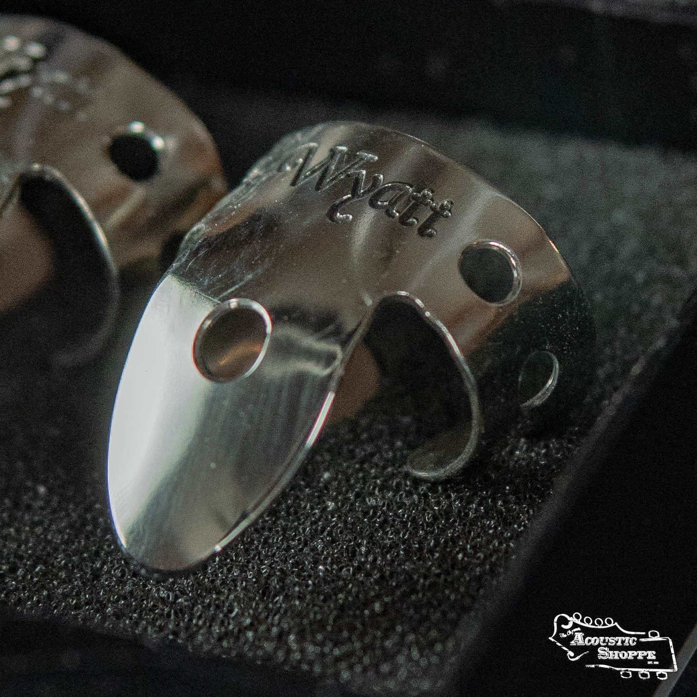 A close-up of a shiny Wyatt Custom Banjos Finger Pick with the “Wyatt Custom Banjos” logo engraved, resting on a textured black surface. Other Wyatt Custom Banjos finger picks appear partially in the background.