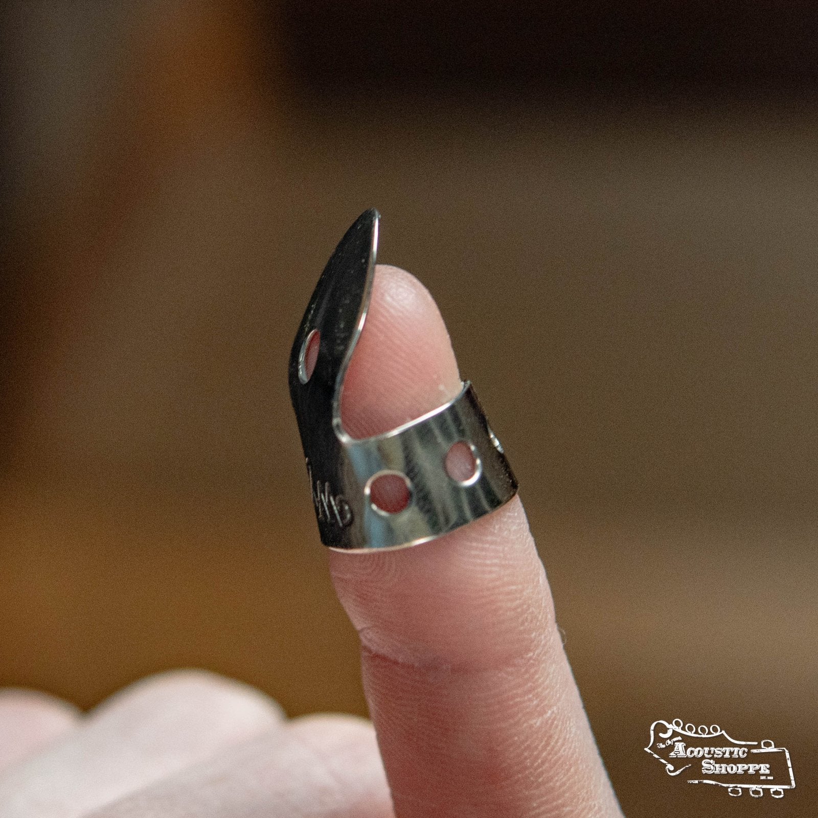 A close-up of a finger wearing a Wyatt Custom Banjos Finger Pick by Wyatt Custom Banjos, made from German Nickel Silver with holes and a curved tip, commonly used for playing stringed instruments like the banjo or guitar.