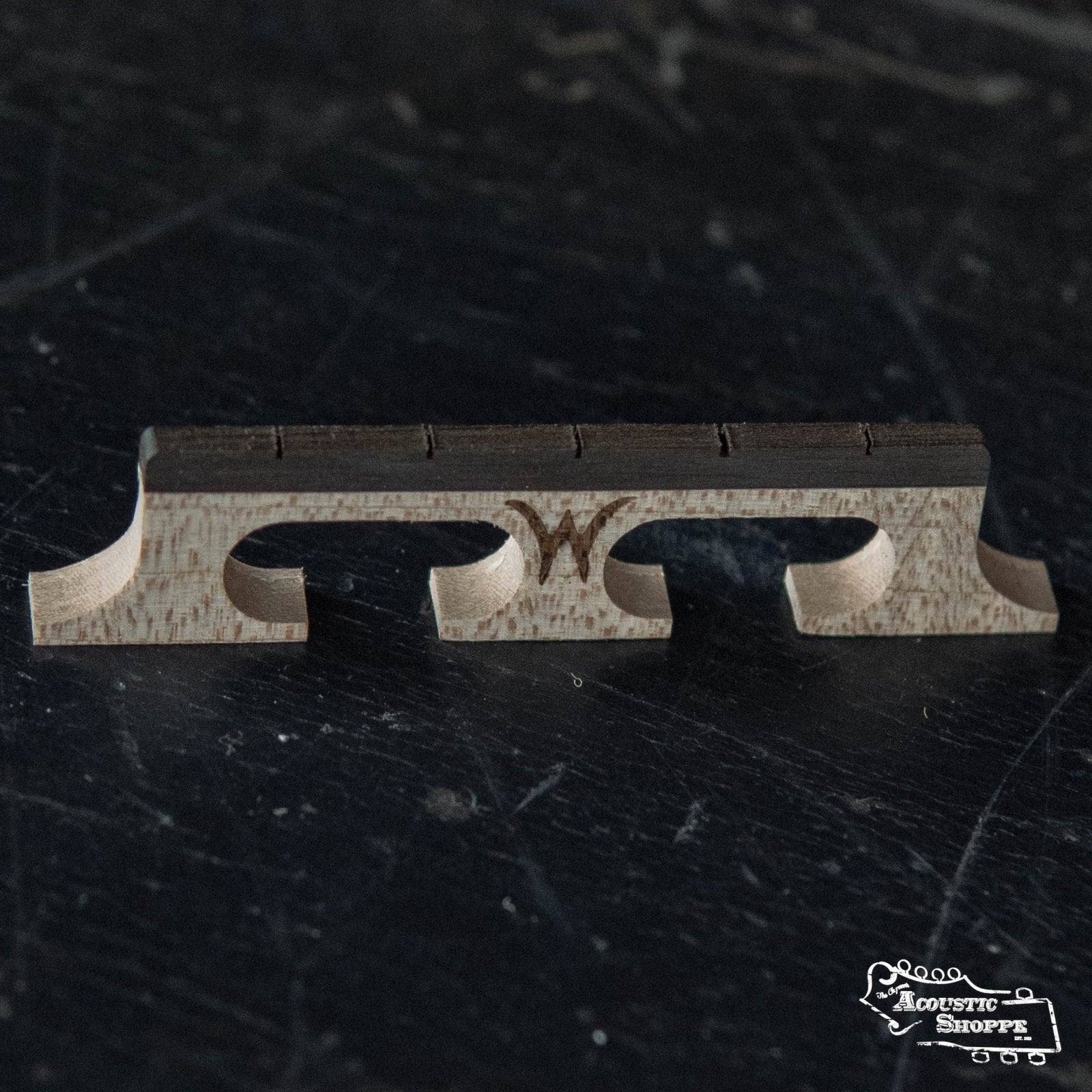 A close-up of the Wyatt Custom Banjos Red Maple 5/8" Banjo Bridge with carved cutouts and a "W" at the center rests on a dark surface, with the Acoustic Shoppe logo shown in the bottom right corner.