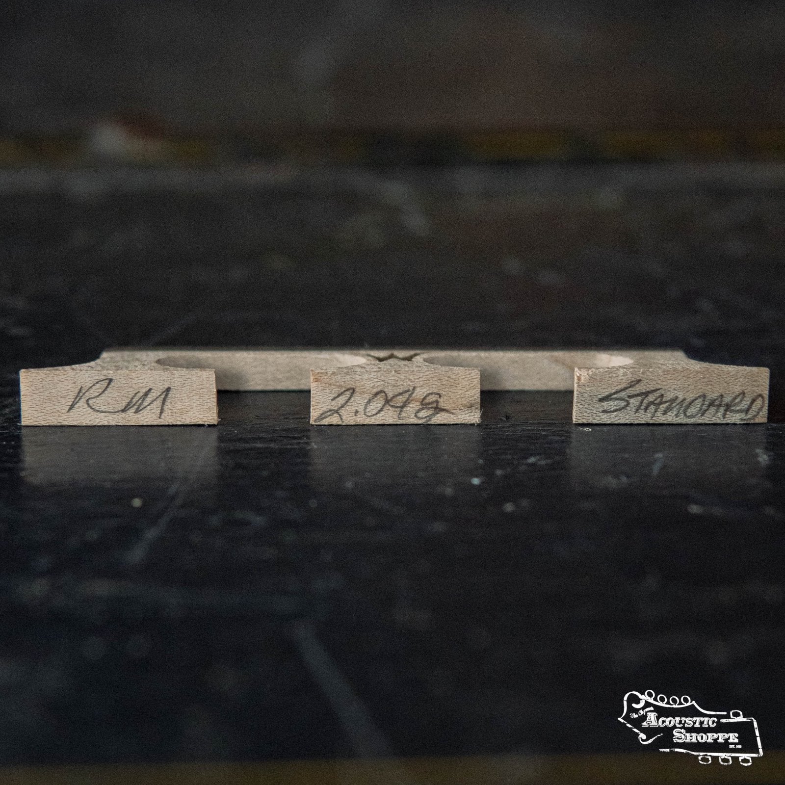 Three small, labeled wood pieces stand upright on a dark surface, showcasing Wyatt Custom Banjos Red Maple 5/8" Banjo Bridge - Standard Spacing by Wyatt Custom Banjos. The Acoustic Shoppe logo appears in the lower right corner.