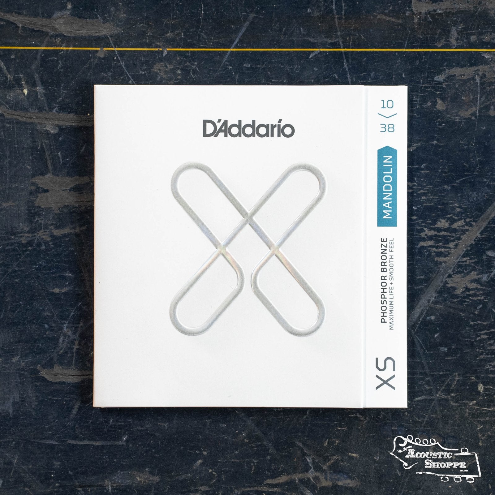 A pack of DAddario 10-38 Light, XS Phosphor Bronze Mandolin Strings sits on a dark, scratched surface; its white minimalist packaging displays a silver string pattern and blue label denoting the gauge and material.