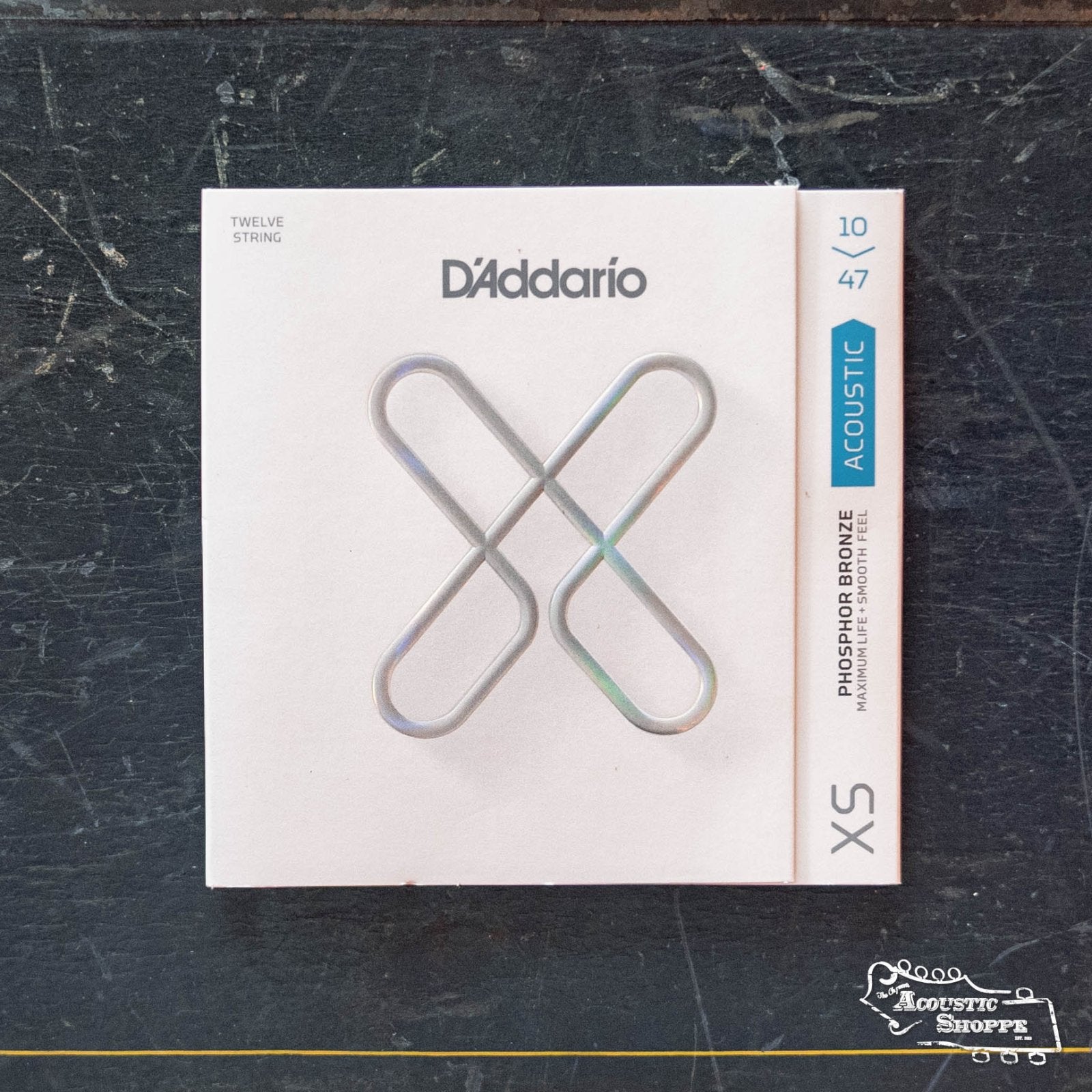 A pack of D'Addario XSAPB1047-12 10-47 Light 12-String XS Phosphor Bronze Coated Acoustic Guitar Strings is displayed on a dark, textured surface with the Acoustic Shoppe logo in the lower right corner.