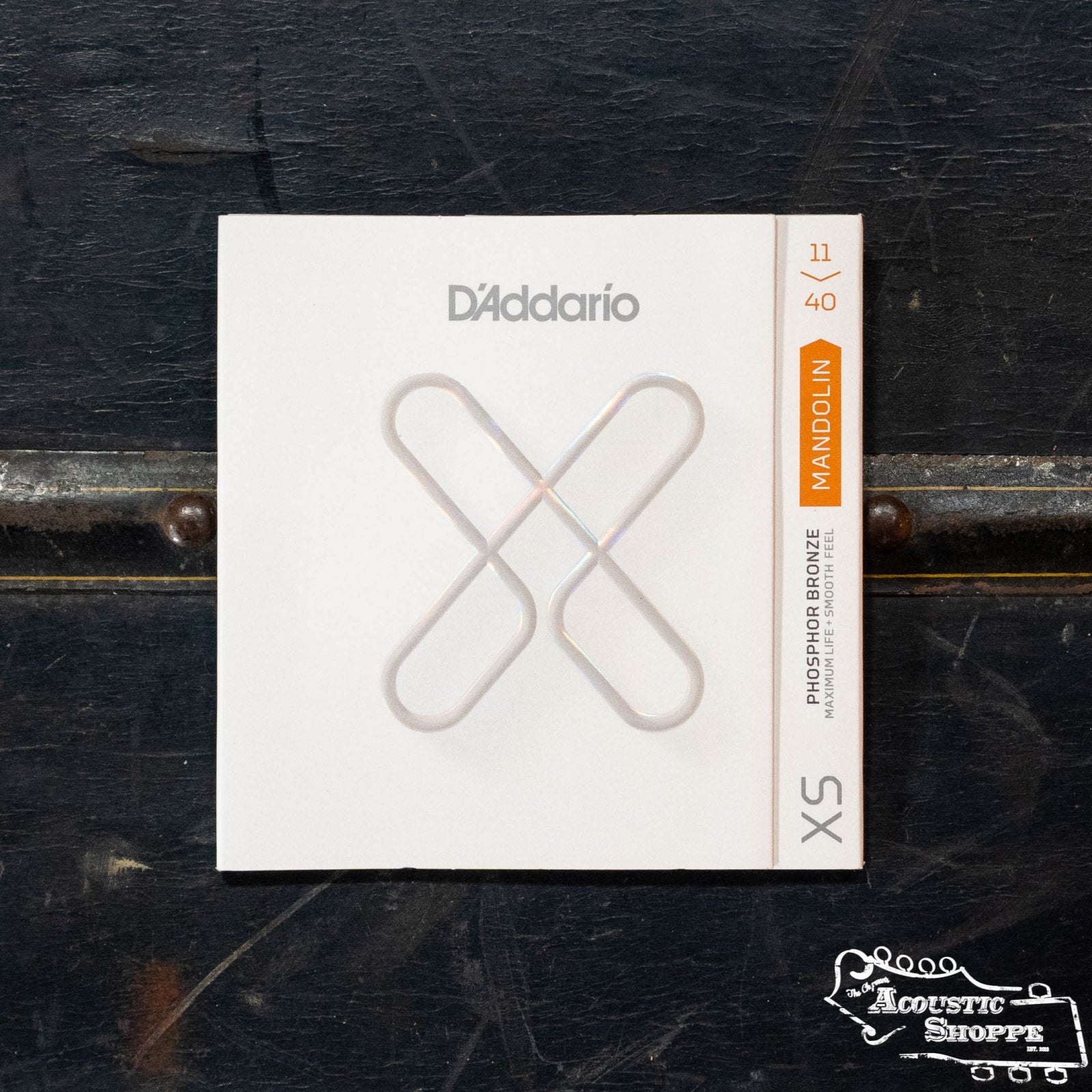 A pack of D’Addario XSM1140 Mandolin Phosphor Bronze Strings (Medium, 11-40) is on a dark wooden surface. The white packaging shows a metallic X, orange mandolin label, and “The Acoustic Shoppe” logo in the corner.
