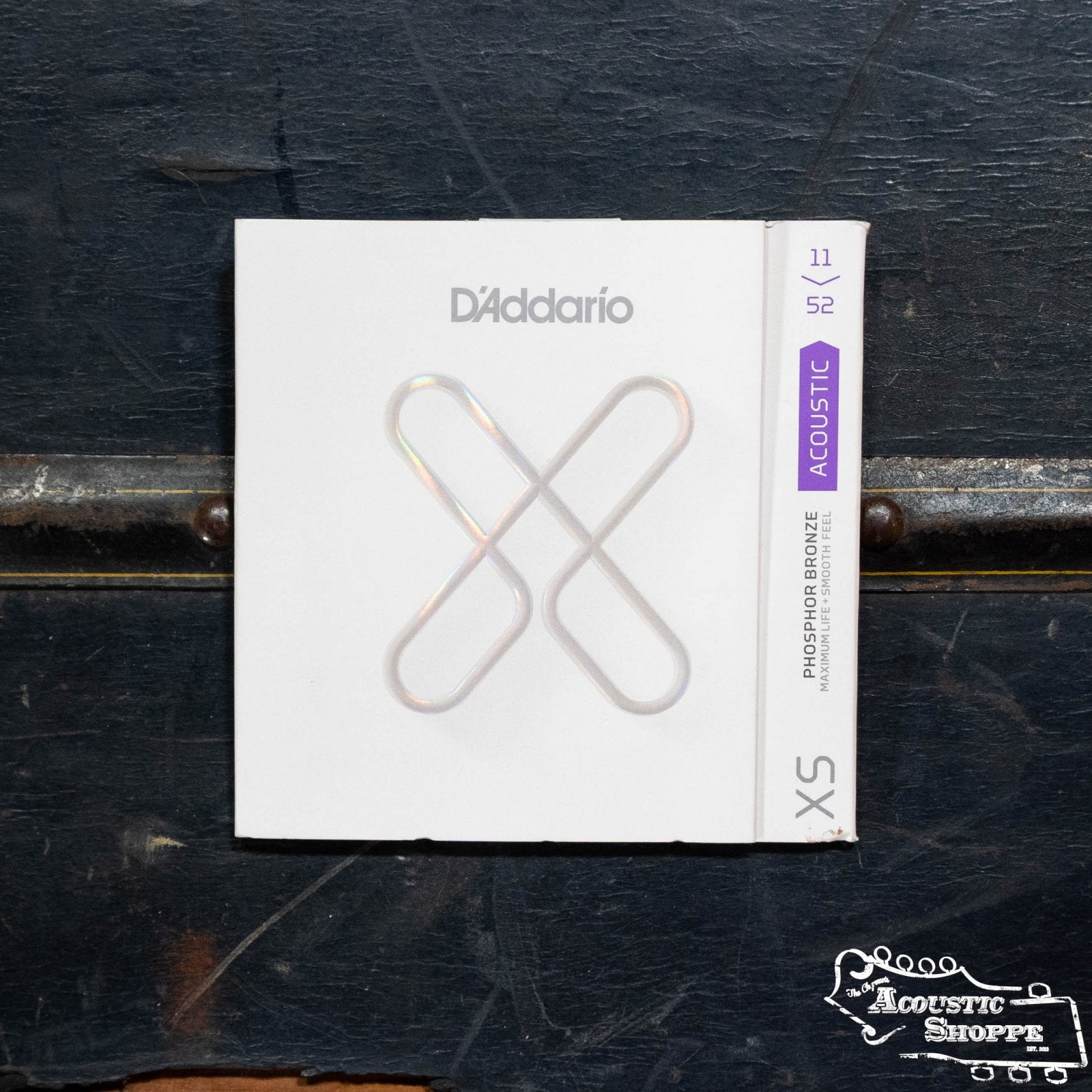 A pack of D'Addario XSAPB1152 XS Custom Light Phosphor Bronze guitar strings (11-52 gauge) sits on a dark rustic surface, with The Acoustic Shoppe logo in the bottom right corner.