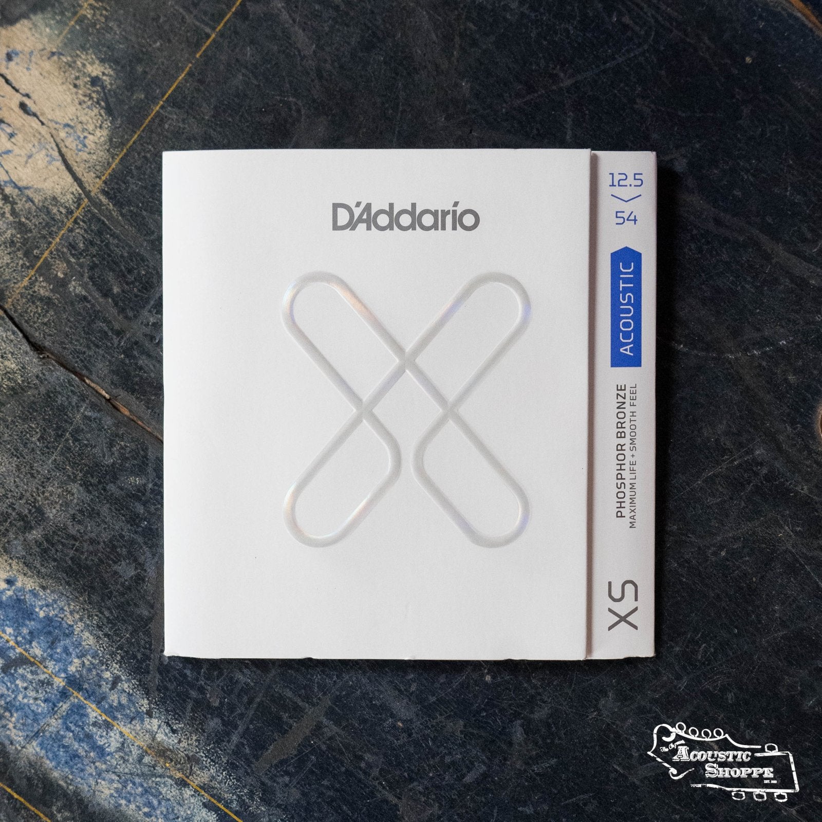 A pack of D'Addario XSAPB12554 XS Phosphor Bronze Acoustic Guitar Strings (Light Plus, 12.5-54) sits on a worn, dark wooden surface; the white packaging with silver and blue accents shows The Acoustic Shoppe logo in the corner.