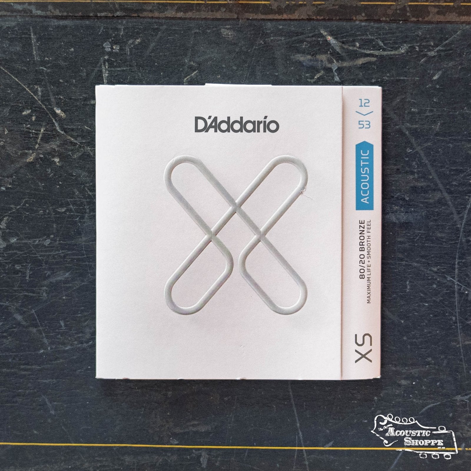 A pack of DAddario XSABR1253 XS 80/20 Bronze Coated Acoustic Guitar Strings, 12-53 Light gauge, rests on a dark, worn surface. The white packaging features a metallic X design with blue text in the corner.