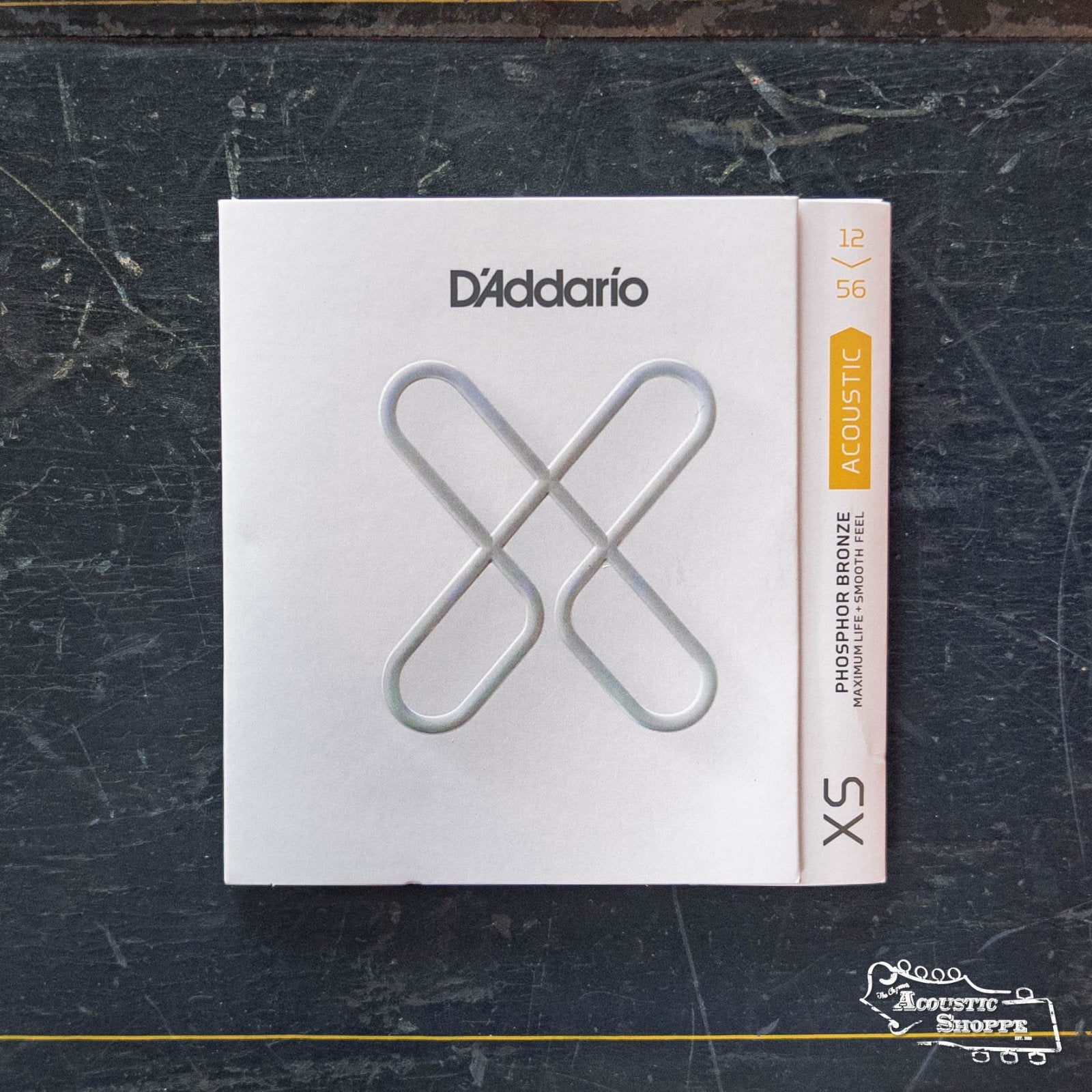 A pack of D'Addario XSAPB1256 XS Light Top/Medium Bottom Phosphor Bronze Guitar Strings, with distinctive white packaging and silver X design, rests on a dark textured surface featuring the Acoustic Strings Shop logo.