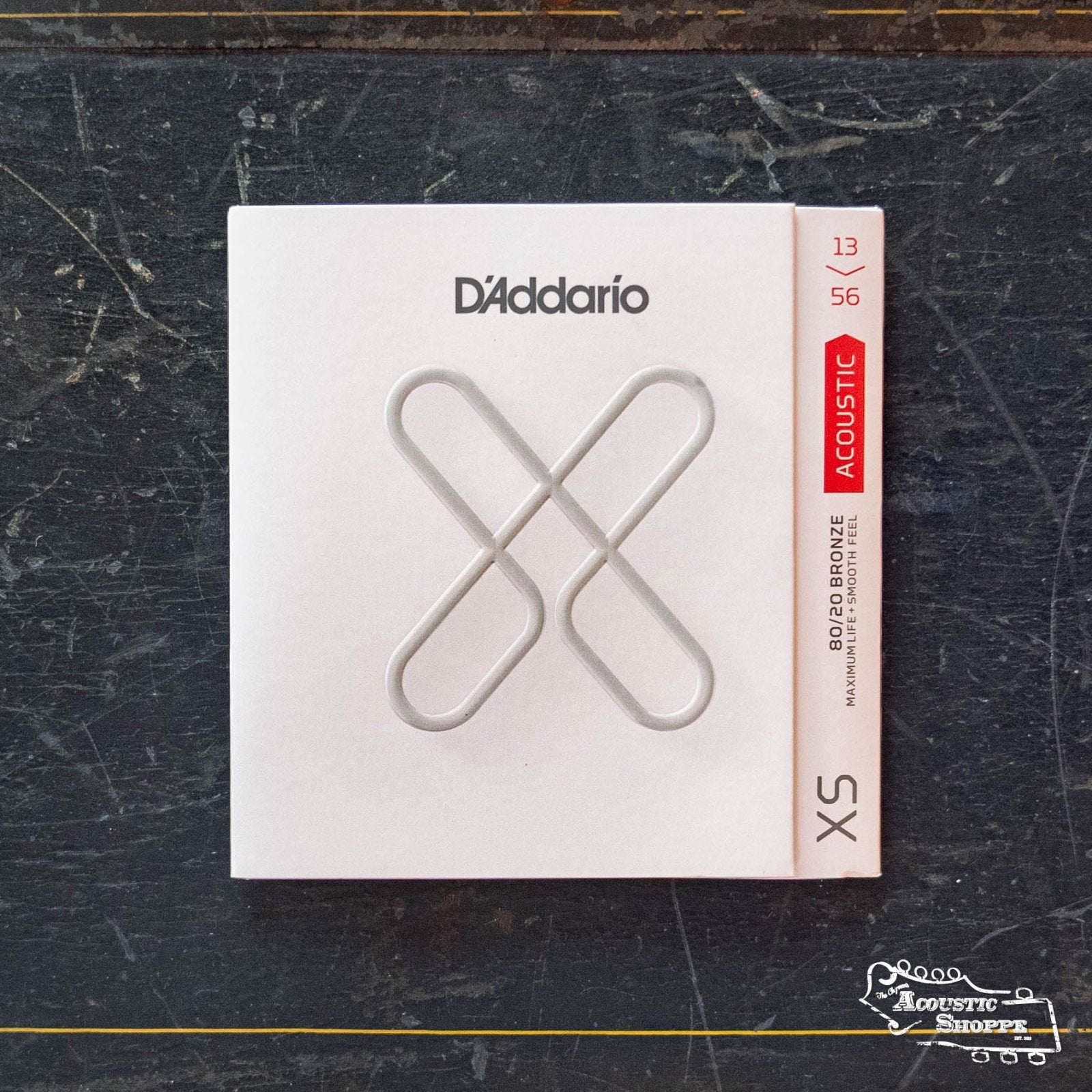 A package of DAddario XSABR1356 XS 80/20 Bronze Coated Acoustic Guitar Strings (13-56 Medium) is shown on a dark, textured surface. The minimalist white packaging features silver lines forming an abstract X.