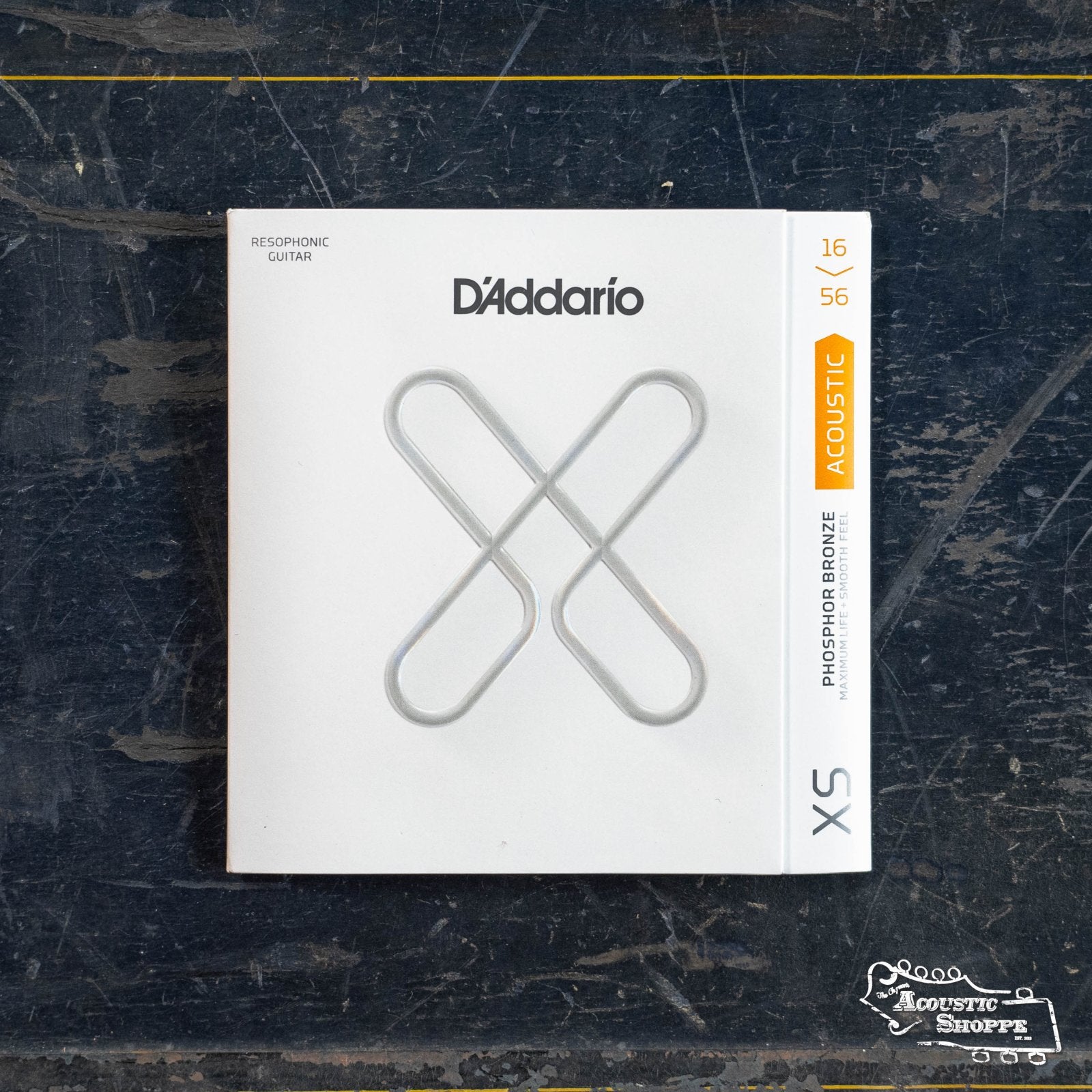 A box of D'Addario XSAPB1656 XS Acoustic Phosphor Bronze resophonic strings, 16-56 gauge, sits on a dark, scratched surface with “The Acoustic Shoppe” logo in the corner.