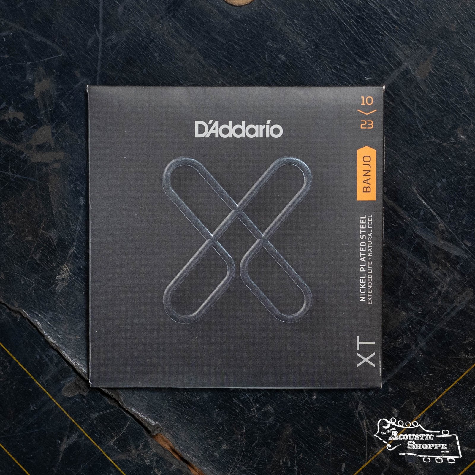 A pack of DAddario XTJ1023 XT Nickel Plated Steel Banjo Strings (medium, 10-23) rests on a textured dark surface. The black, silver X packaging highlights the corrosion-resistant quality of these premium DAddario strings.