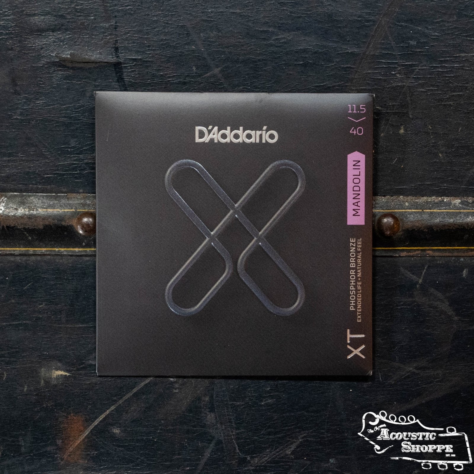 A pack of D’Addario XTM11540 Phosphor Bronze Custom Medium Mandolin Strings rests on a dark wooden surface; the black packaging displays a stylized X and product details in white and purple text.