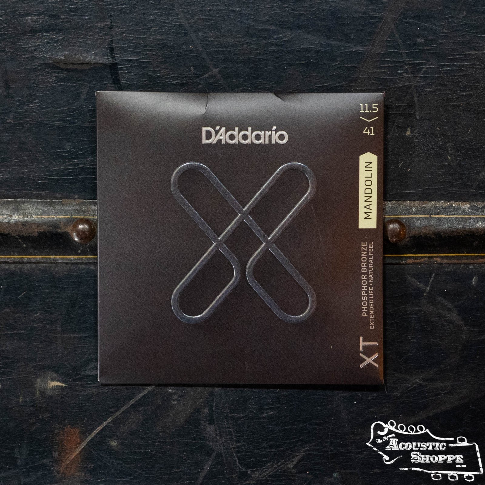 A box of DAddario XTM11541 Phosphor Bronze Medium/Heavy Mandolin strings with extended lifespan sits on a dark wooden surface. The mostly black DAddario box shows a metallic X and a yellow Mandolin label.