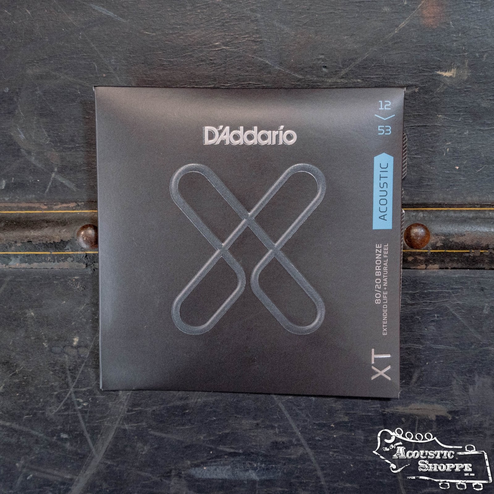 A pack of D'Addario XTABR1253 80/20 Bronze Light Acoustic Guitar Strings (12-53 gauge) with a black, silver X design and blue accent, offering extended lifespan, displayed on a dark textured background.