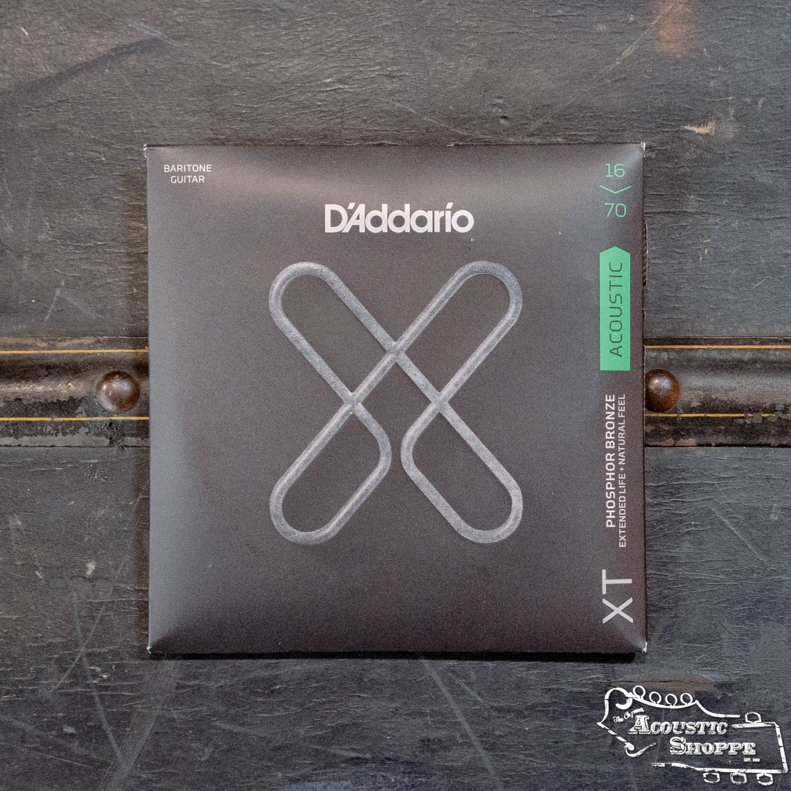 A pack of DAddario XTAPB1670 Phosphor Bronze Coated Acoustic Baritone Guitar Strings, Medium 16-70, is displayed on a dark, textured surface with The Acoustic Shoppe logo in the bottom right corner.