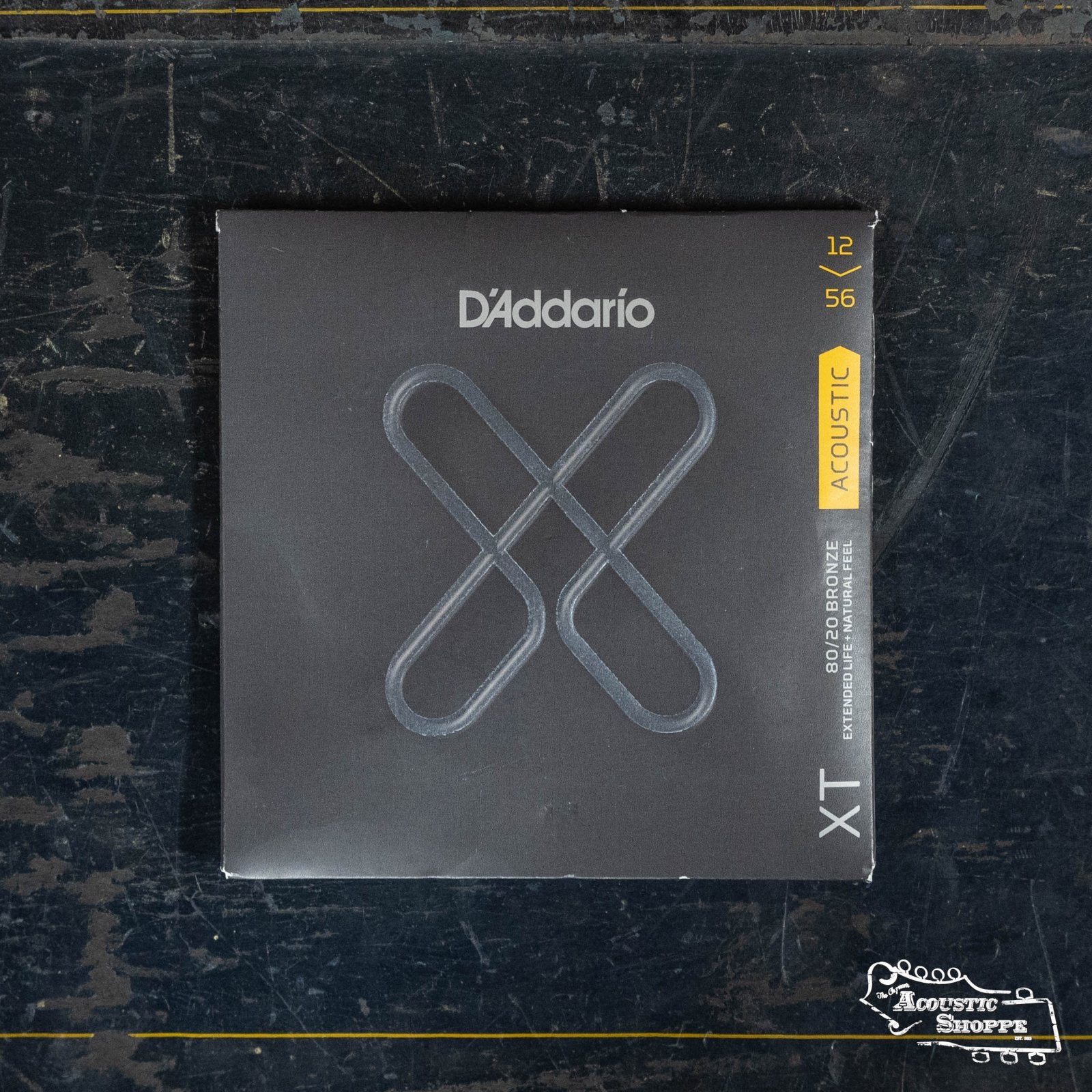 A pack of D'Addario XTABR1256 XT 80/20 Bronze Acoustic Guitar Strings (Light Top/Medium, 12-56) rests on a scratched dark surface. Its black packaging features a silver X and yellow acoustic label, boasting superior XT corrosion-resistant coating.