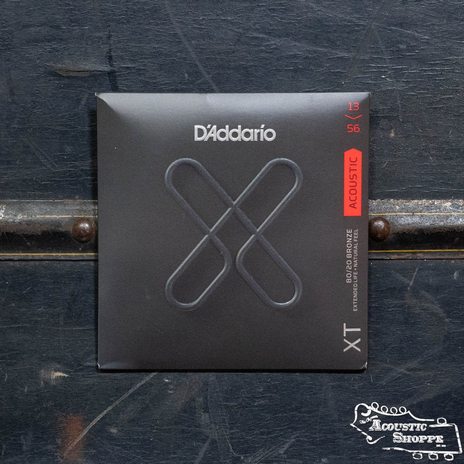 The D'Addario XTABR1356 80/20 Bronze Medium Acoustic Guitar Set offers extended lifespan, black packaging with a bold X design, polymer coating, and is labeled 13-56. The Acoustic Shoppe logo is at the bottom right corner.