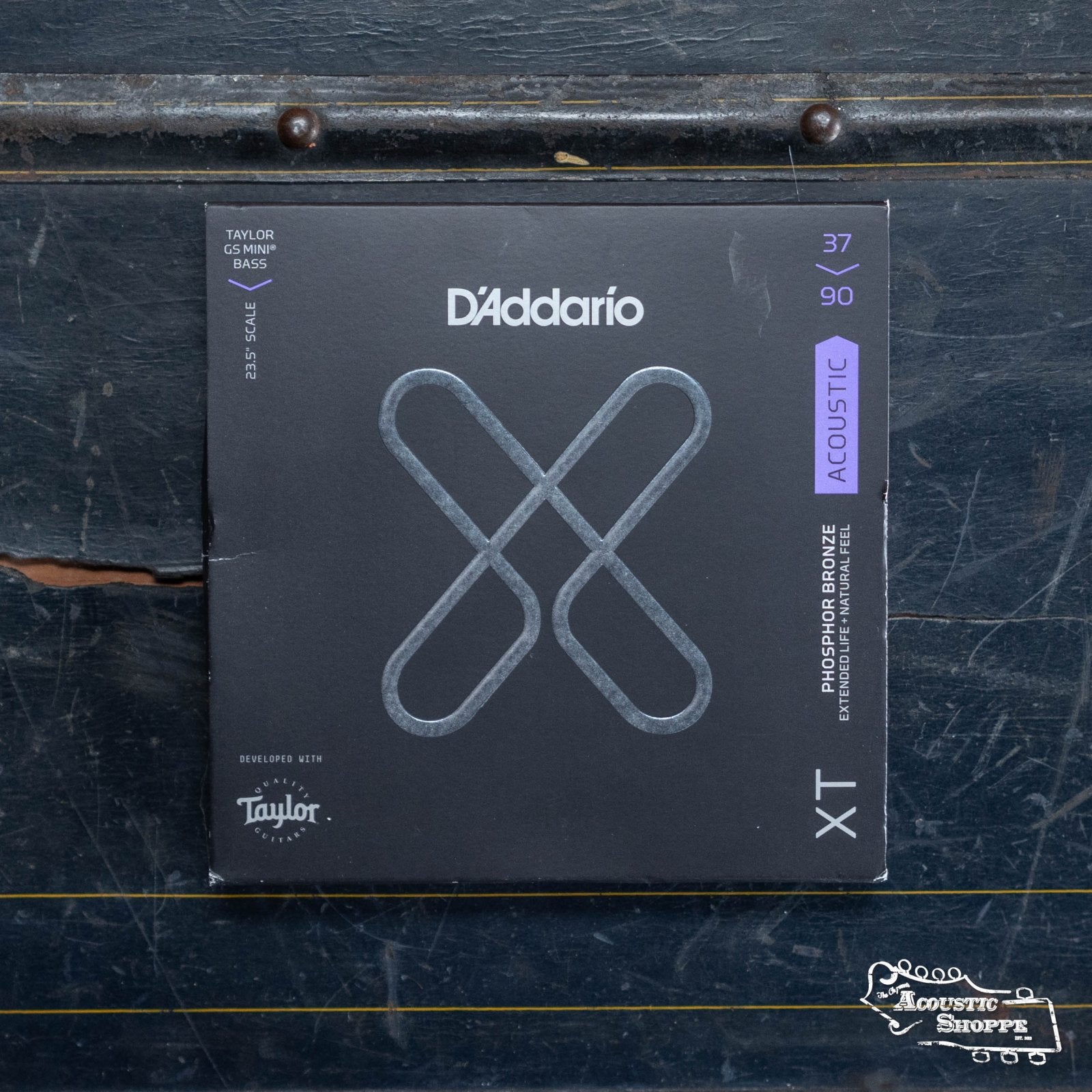 A box of D'Addario XTB3790GS Phosphor Bronze Custom Light Taylor GS Mini Coated Acoustic Bass Strings (37-90) features a gray background with blue and white text, a large X on the front, and The Acoustic Shoppe logo in the lower right.