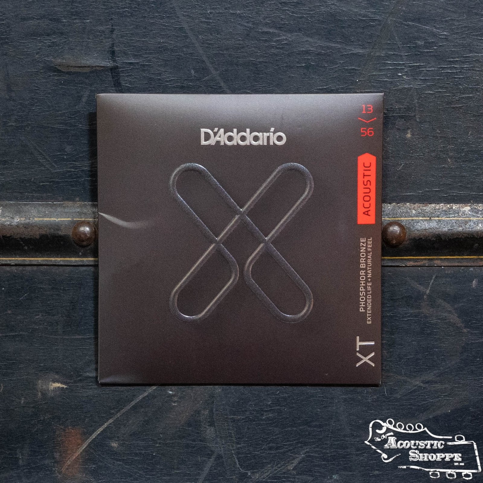 A package of DAddario XTAPB1356 Phosphor Bronze Medium Acoustic Guitar Strings rests on a dark wooden surface, its black packaging displaying a bold “X” design and an orange label showing the 13-56 gauge.