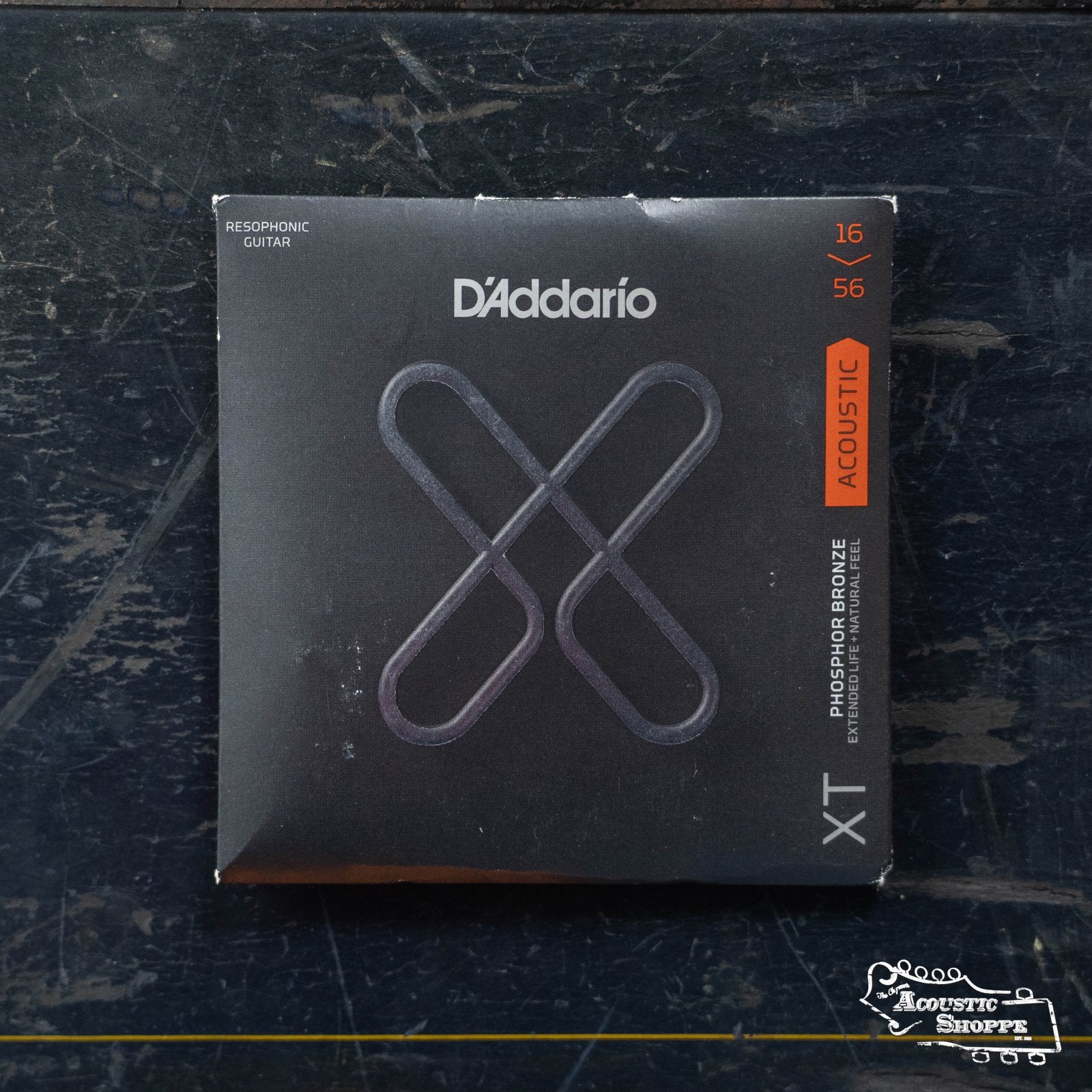A pack of DAddario XTAPB1656 XT Acoustic Phosphor Bronze Resophonic guitar strings (16–56 medium gauge) sits on a scratched dark surface, featuring black packaging with silver and orange accents and a large stylized X.