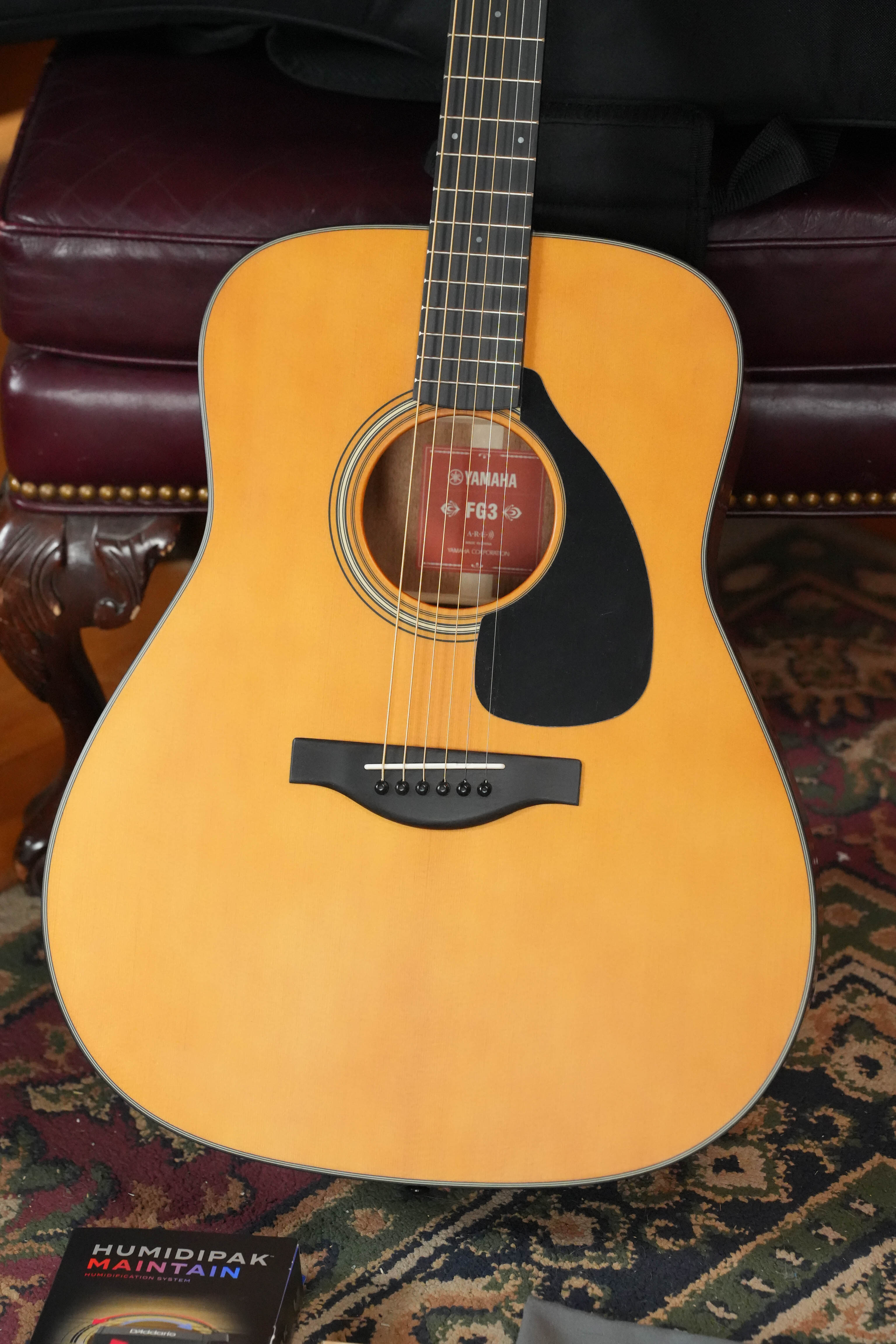 A Yamaha FG3 Red Label Dreadnought Acoustic Guitar #1160 with a solid Sitka spruce top and natural finish rests on an ornate rug before a dark red leather chair, with a Humidipak Maintain packet in the lower left corner.