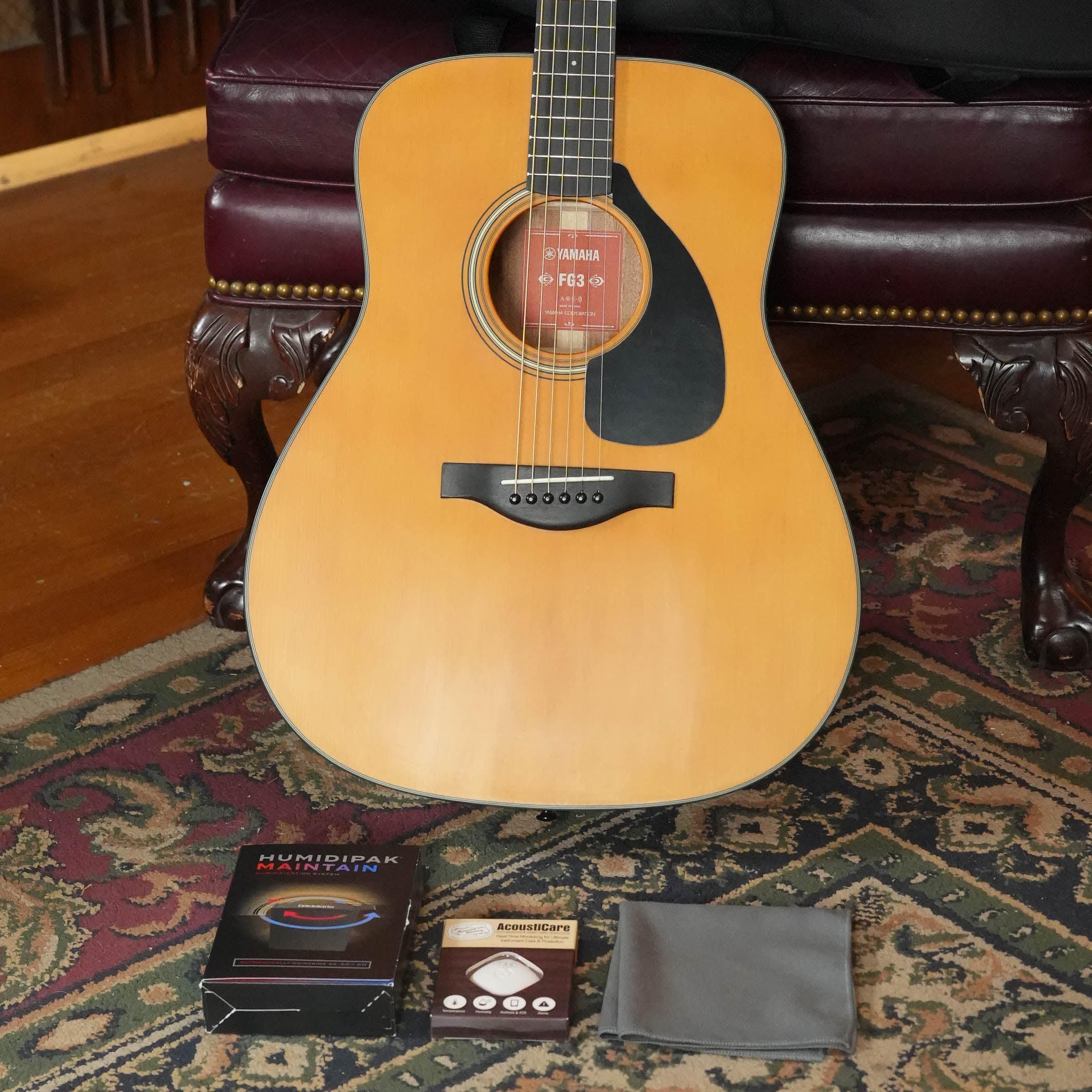 A Yamaha FG3 Red Label Dreadnought Acoustic Guitar #1210 by Yamaha stands upright on a patterned rug, with guitar strings, a maintenance kit, and a gray cleaning cloth in front, leaning against a dark upholstered chair.