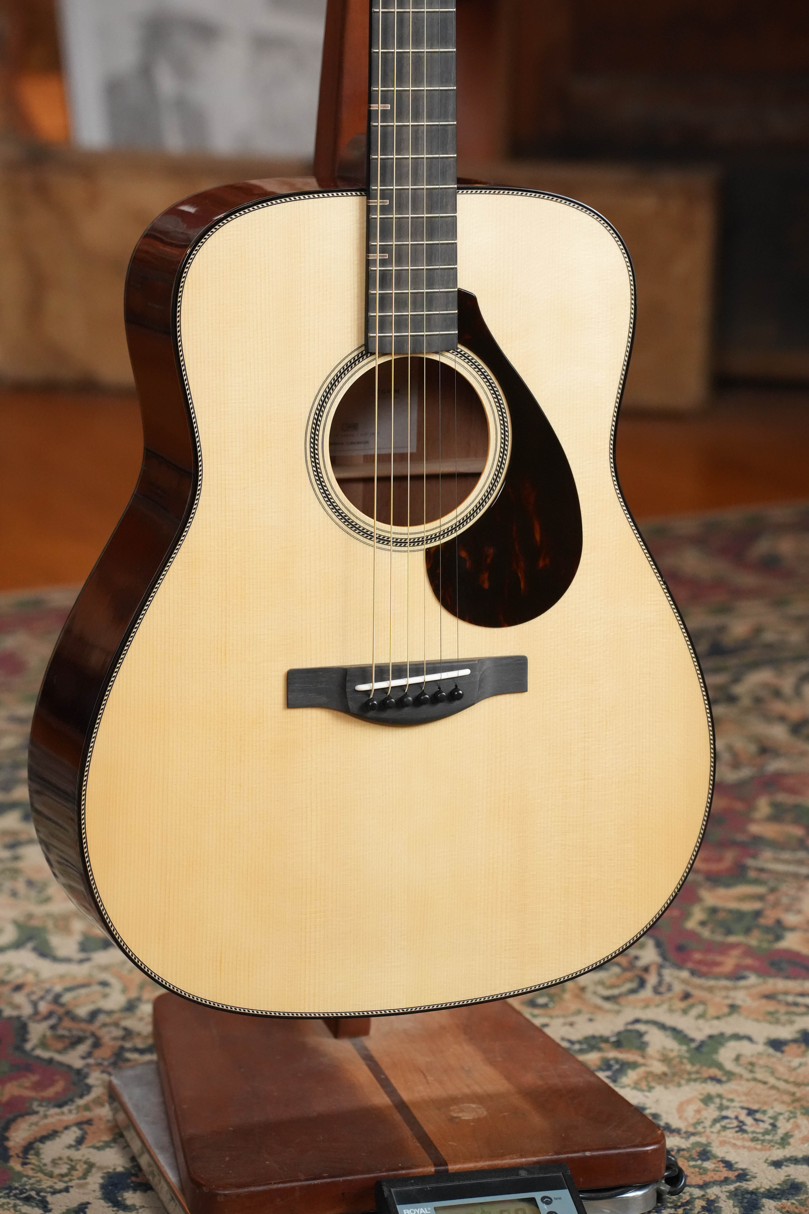 A close-up of the Yamaha FG9 M Adirondack/Mahogany Dreadnought Acoustic Guitar #112A by Yamaha, featuring an Adirondack spruce top, dark mahogany sides, black pickguard, and decorative binding on a wooden stand over a patterned rug.
