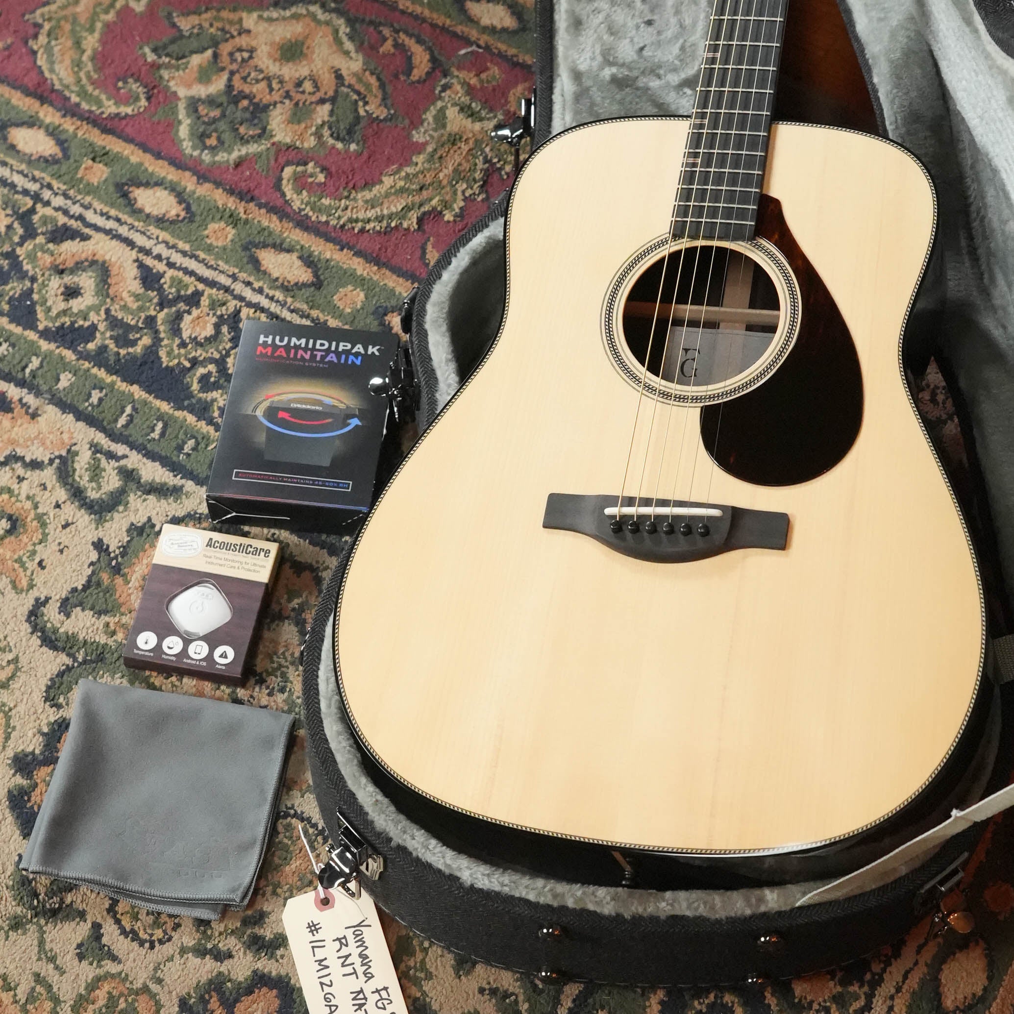 A Yamaha FG9 R NT Natural Finish Dreadnought Acoustic Guitar rests in an open case on a patterned rug, with accessories like a cleaning cloth, humidifier pack, extra pick, and a thank-you tag for playing #AlvarezGuitars!.