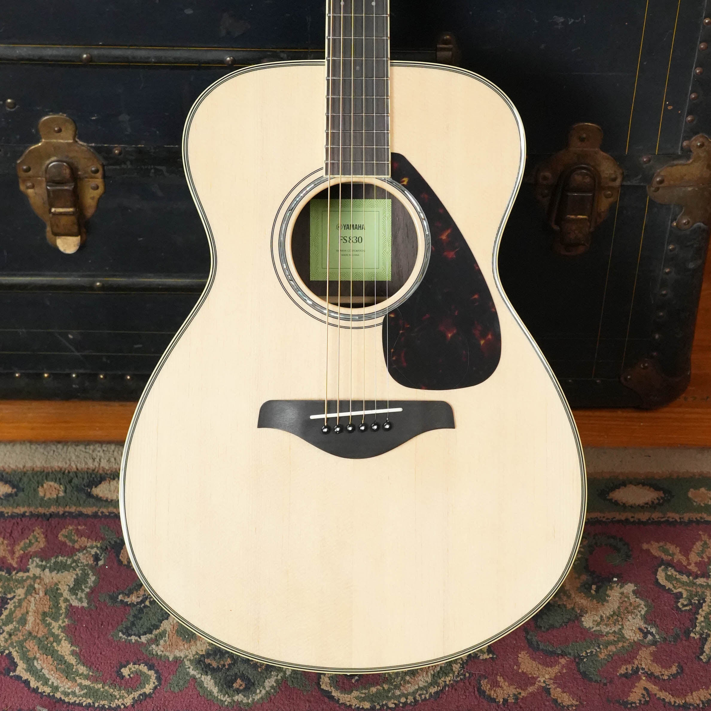 A Yamaha FS830 Concert Acoustic Guitar #1751 by Yamaha, featuring a solid spruce top and dark pickguard, stands upright before a vintage black trunk with part of a patterned rug visible beneath.