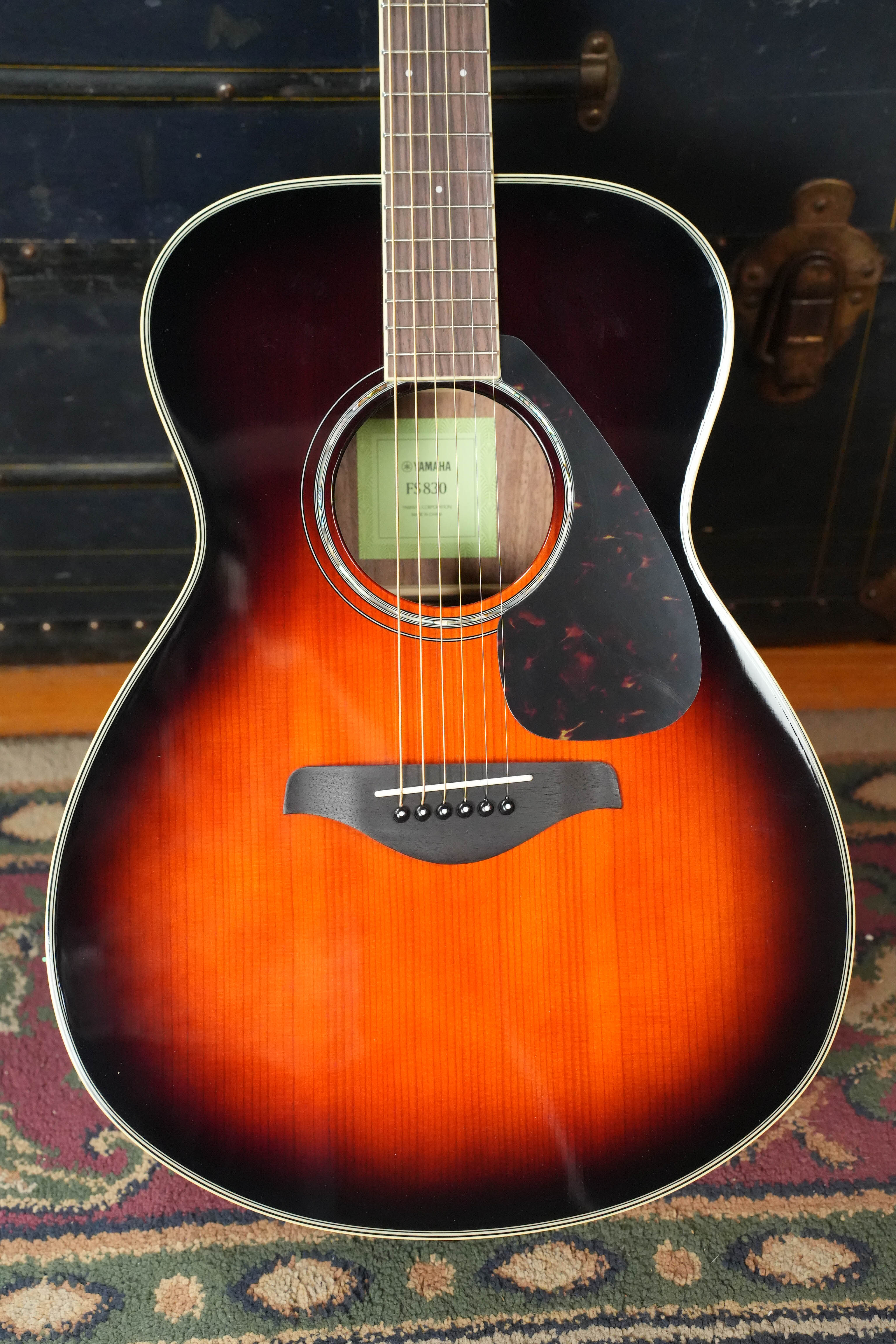 A Yamaha FS830 TBS Tobacco Sunburst Concert Acoustic Guitar (#0129) stands upright on a patterned rug with a dark trunk in the background. The solid spruce top, sunburst finish, and tortoiseshell pickguard complement its classic Yamaha style.