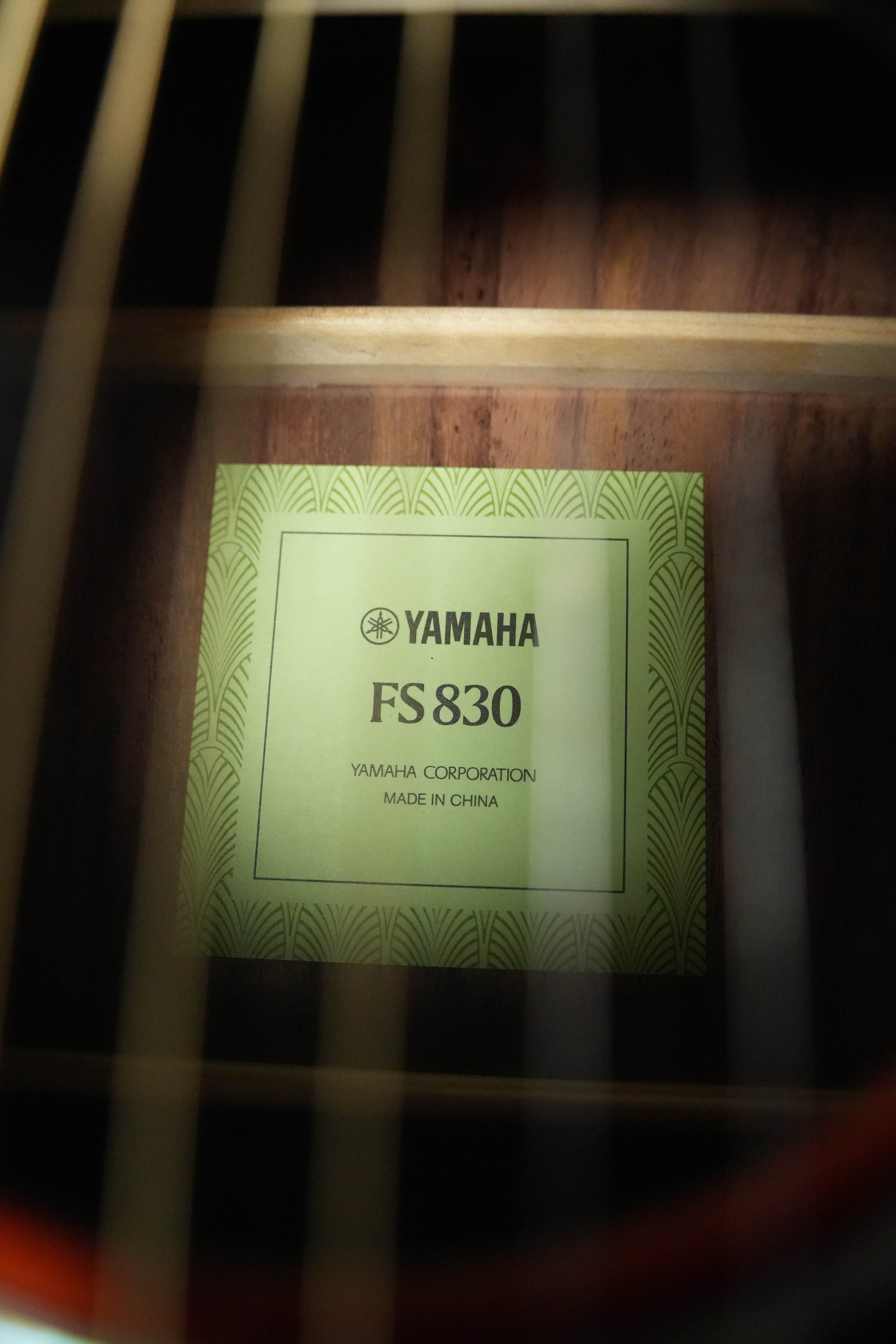 Close-up of a label inside a Yamaha FS830 TBS Tobacco Sunburst Concert Acoustic Guitar #0129, reading “Yamaha,” framed by the guitar strings and wood interior with its solid spruce top subtly visible.