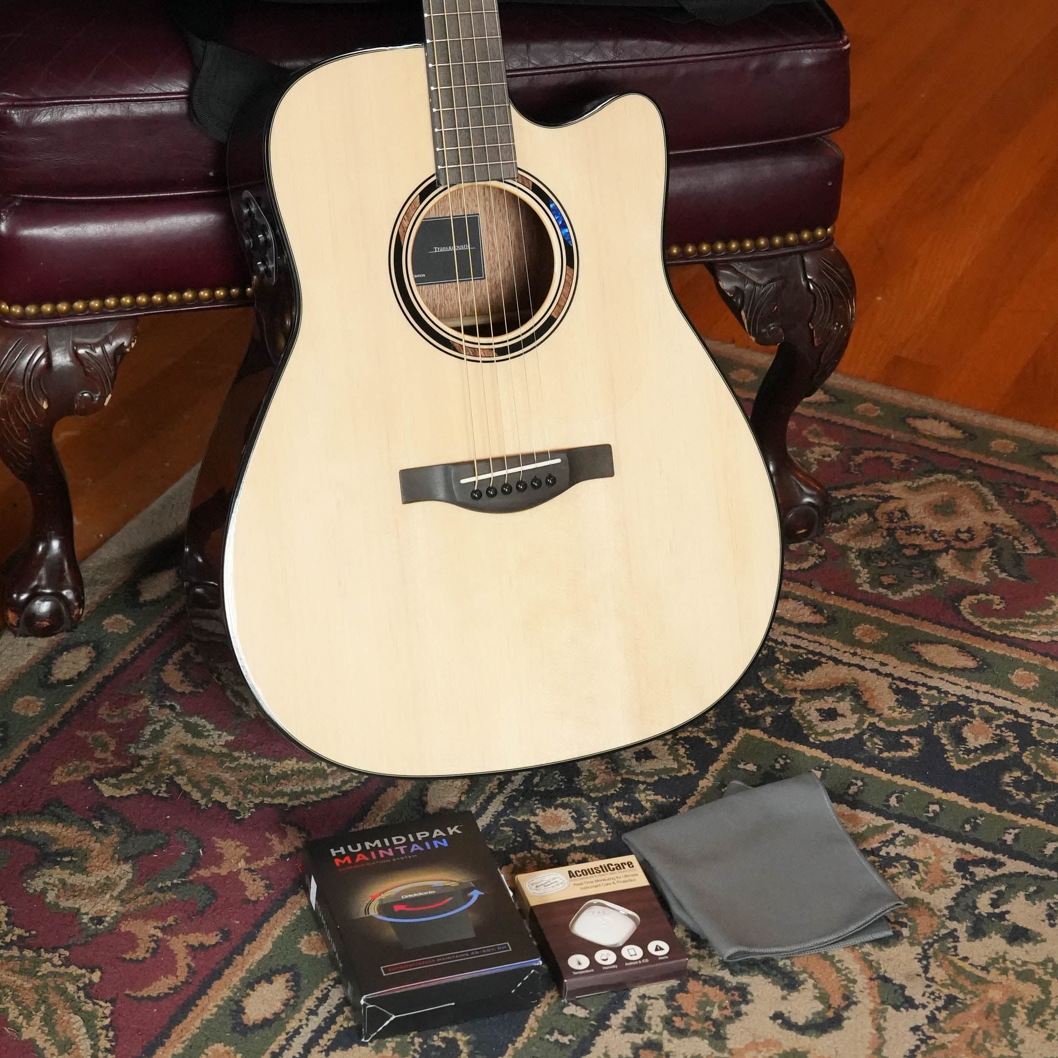 A Yamaha TAG1 C Natural Finish Dreadnought Cutaway Acoustic-Electric Guitar by Yamaha leans against a maroon leather chair on a patterned rug, with a Humidipak kit, small box, and gray cleaning cloth placed in front.