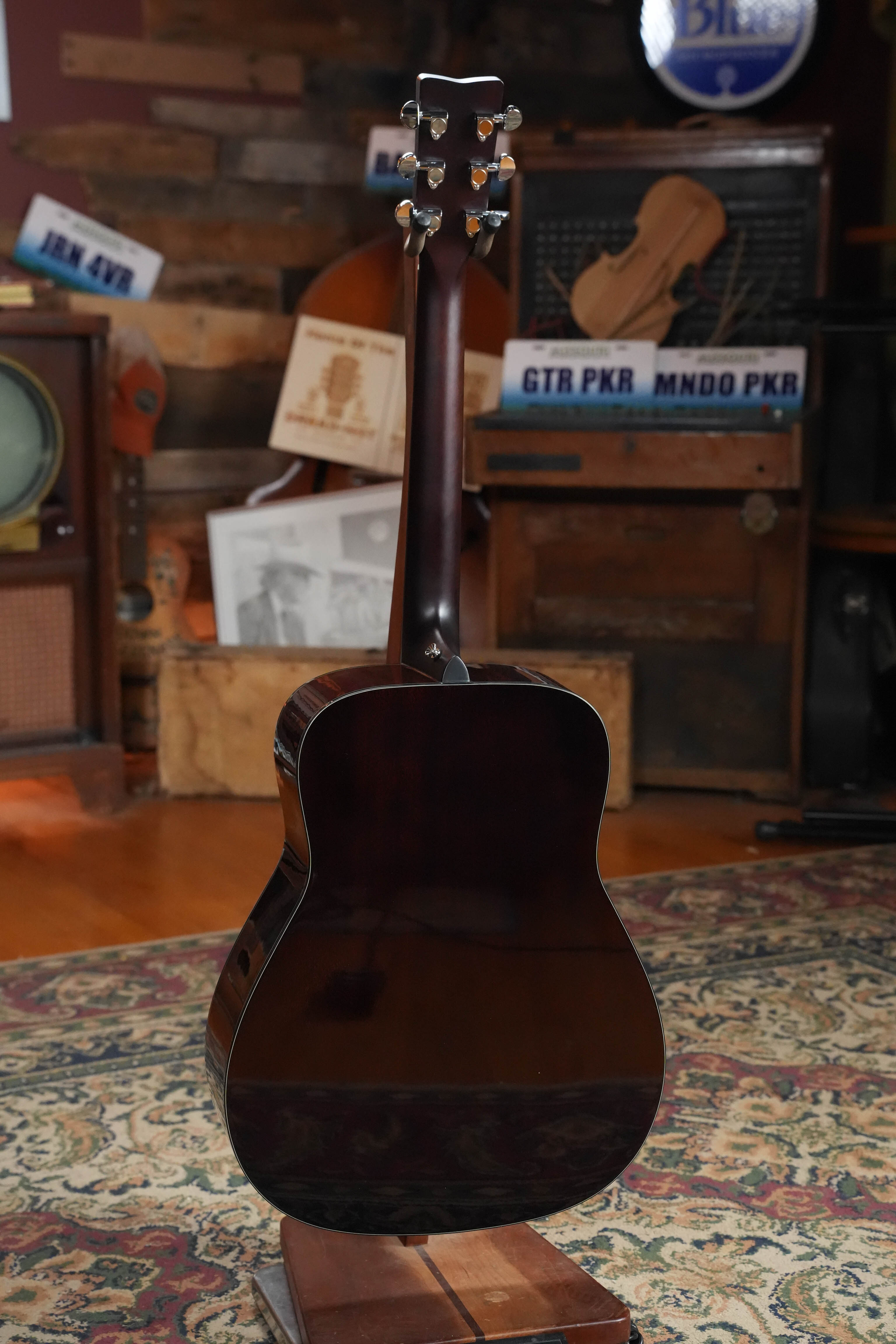 The Yamaha TAG1E TBL Spruce/Mahogany Transacoustic Dreadnought Guitar #1280 by Yamaha stands upright, displaying its back and neck against a backdrop of vintage decor featuring radios, license plates, and musical instruments on shelves.