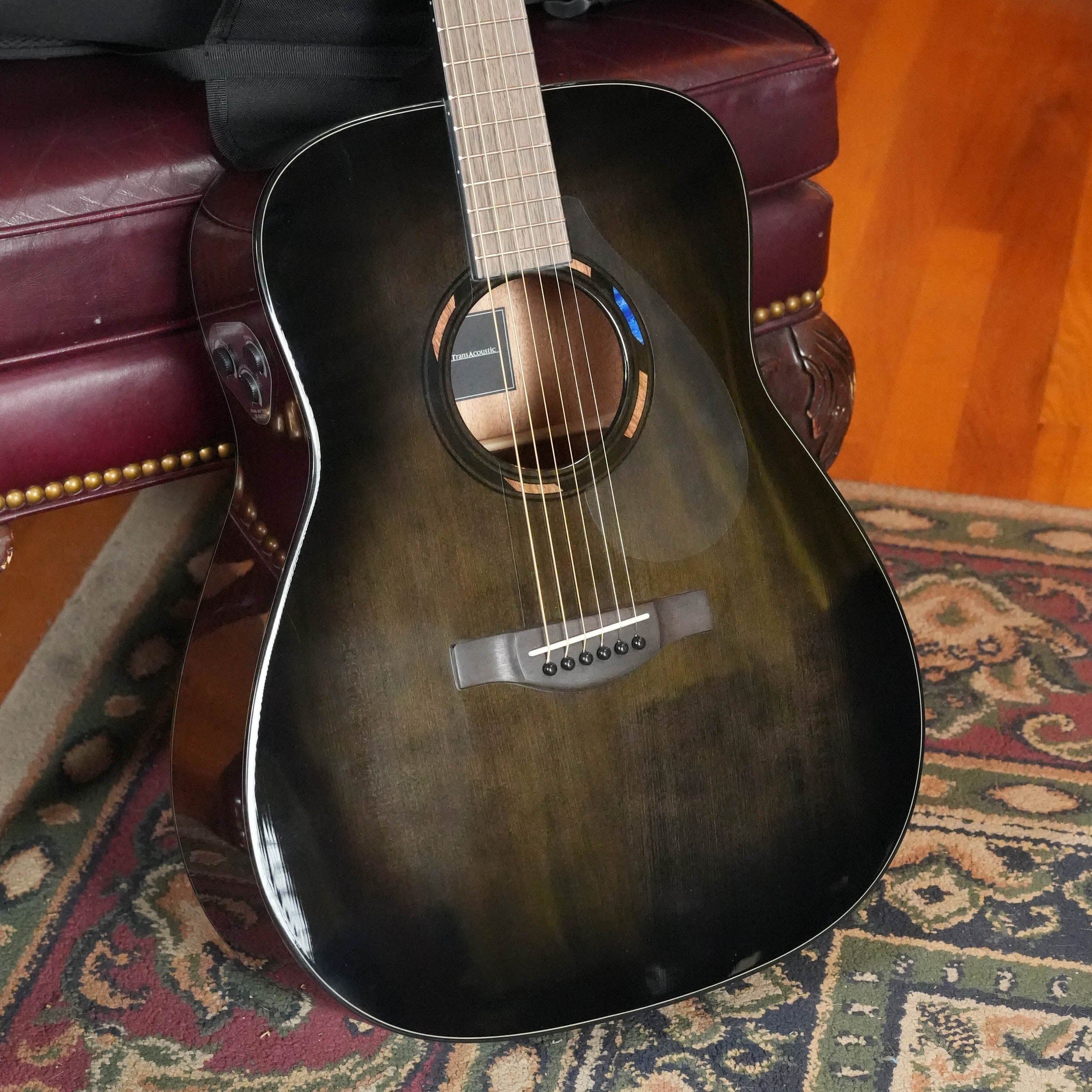 A Yamaha TAG1E TBL TransAcoustic Translucent Black acoustic-electric guitar rests against a maroon leather couch with brass studs, next to a colorful patterned rug on a hardwood floor.