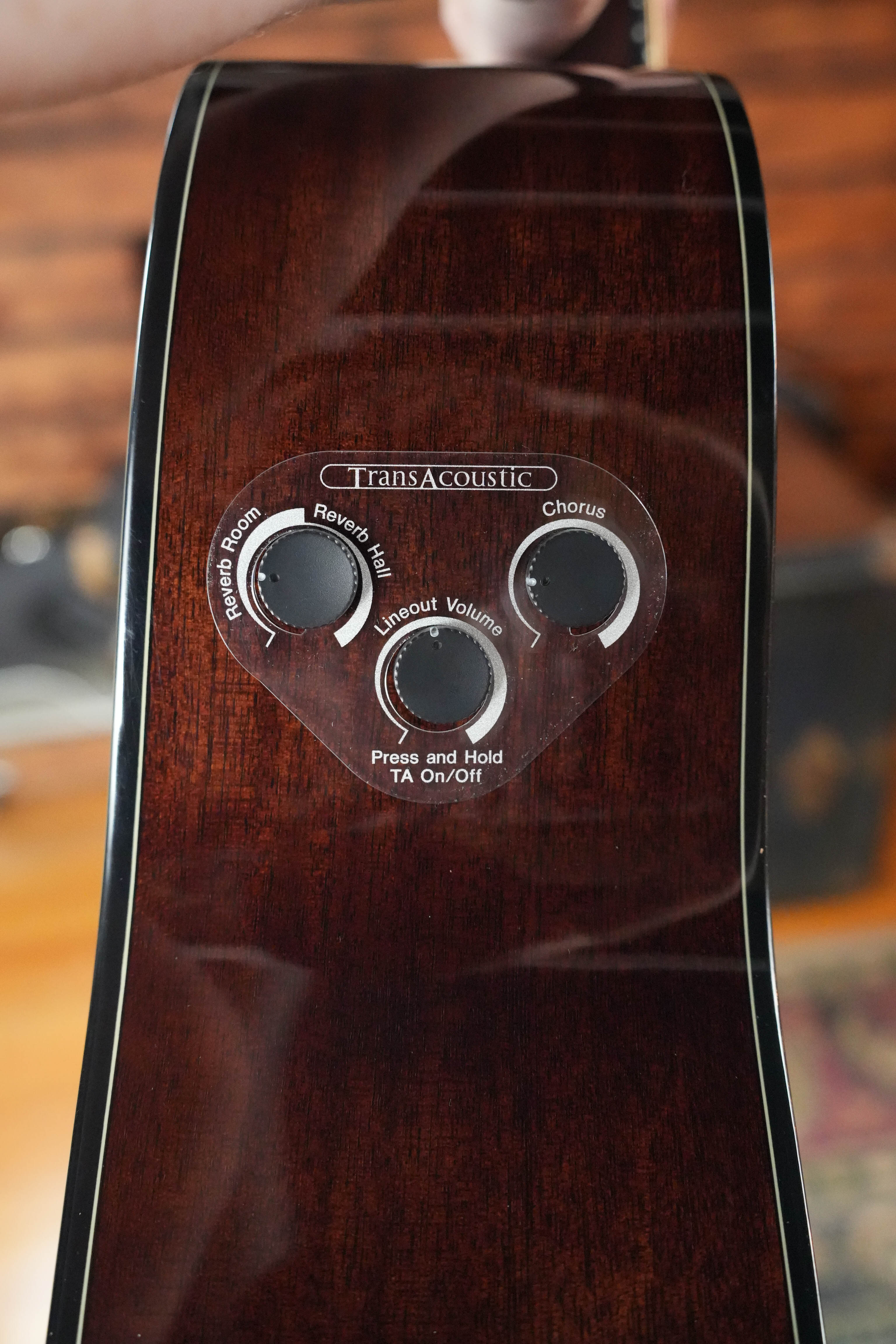 Close-up of the Yamaha TAG1E TBL TransAcoustic (Translucent Black) side panel, highlighting controls for Reverb Room, Reverb Hall, Chorus, Lineout Volume, and TA On/Off—all labeled under the TransAcoustic system.