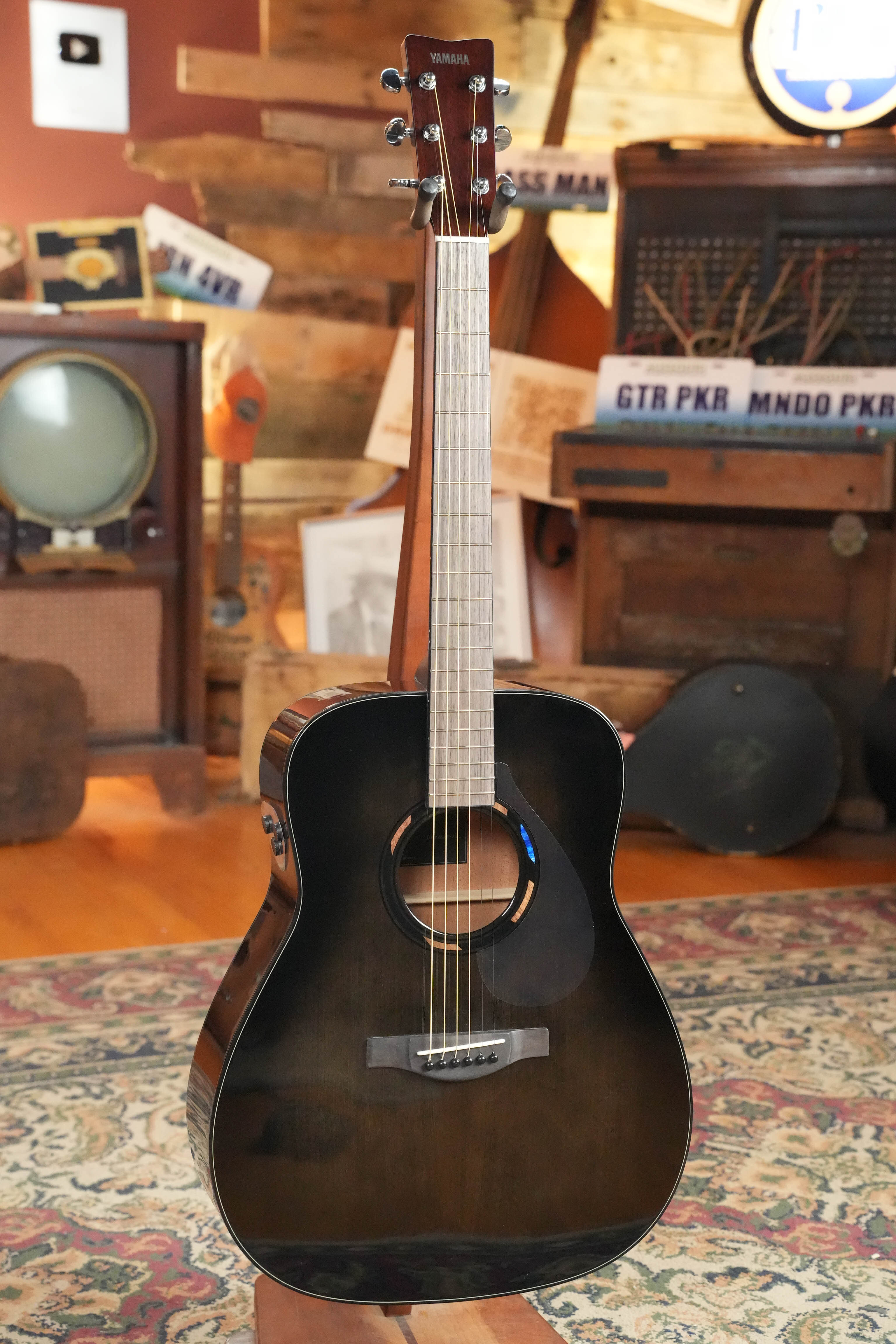 A Yamaha TAG1E TBL TransAcoustic Dreadnought Acoustic-Electric Guitar in Translucent Black (#1164) stands upright on a patterned rug in a vintage-style room with retro decor and musical equipment, featuring the innovative TransAcoustic system.
