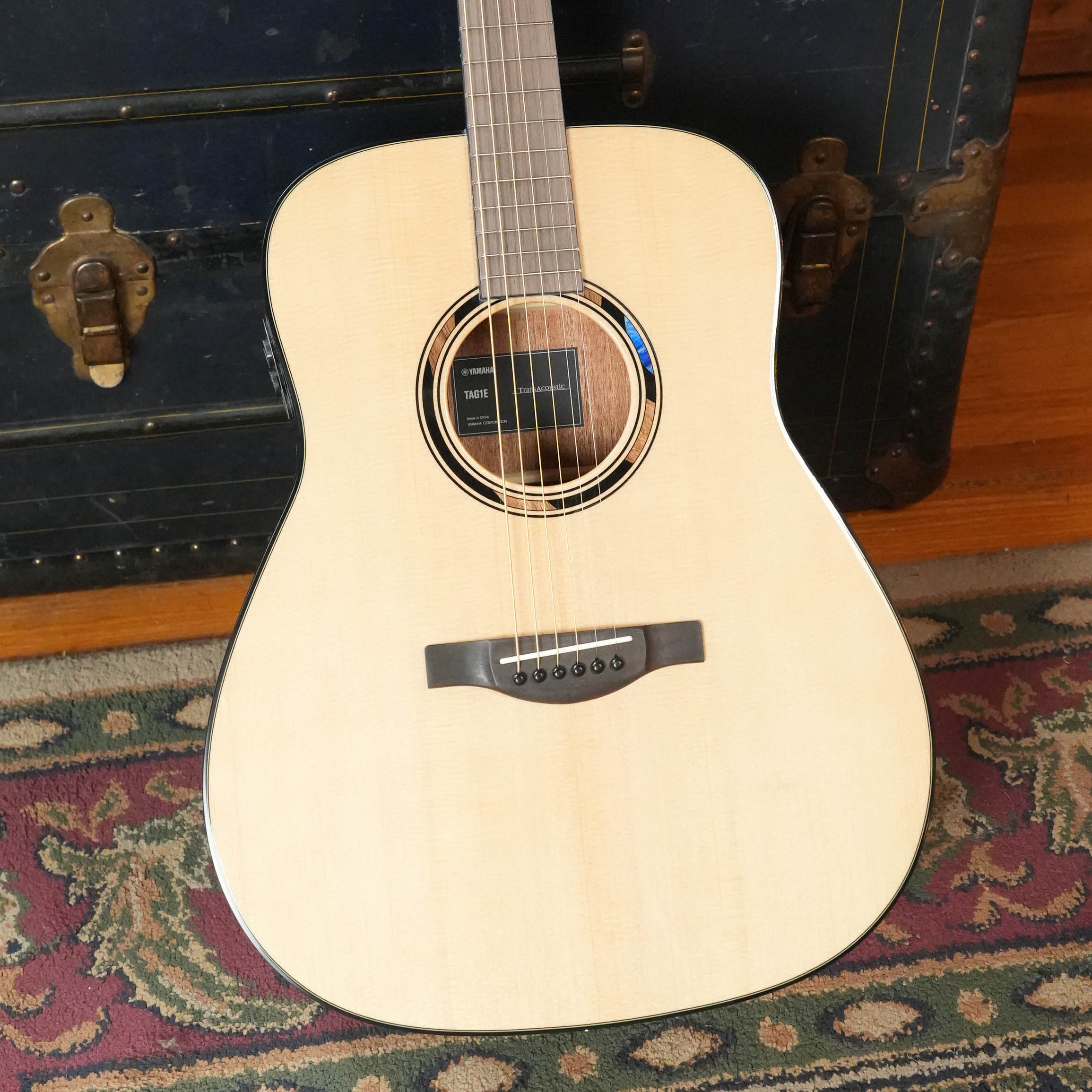 The Yamaha TAS1E Natural Finish Concert Acoustic-Electric Guitar by Yamaha rests upright against a dark antique trunk, with part of a patterned rug visible underneath.