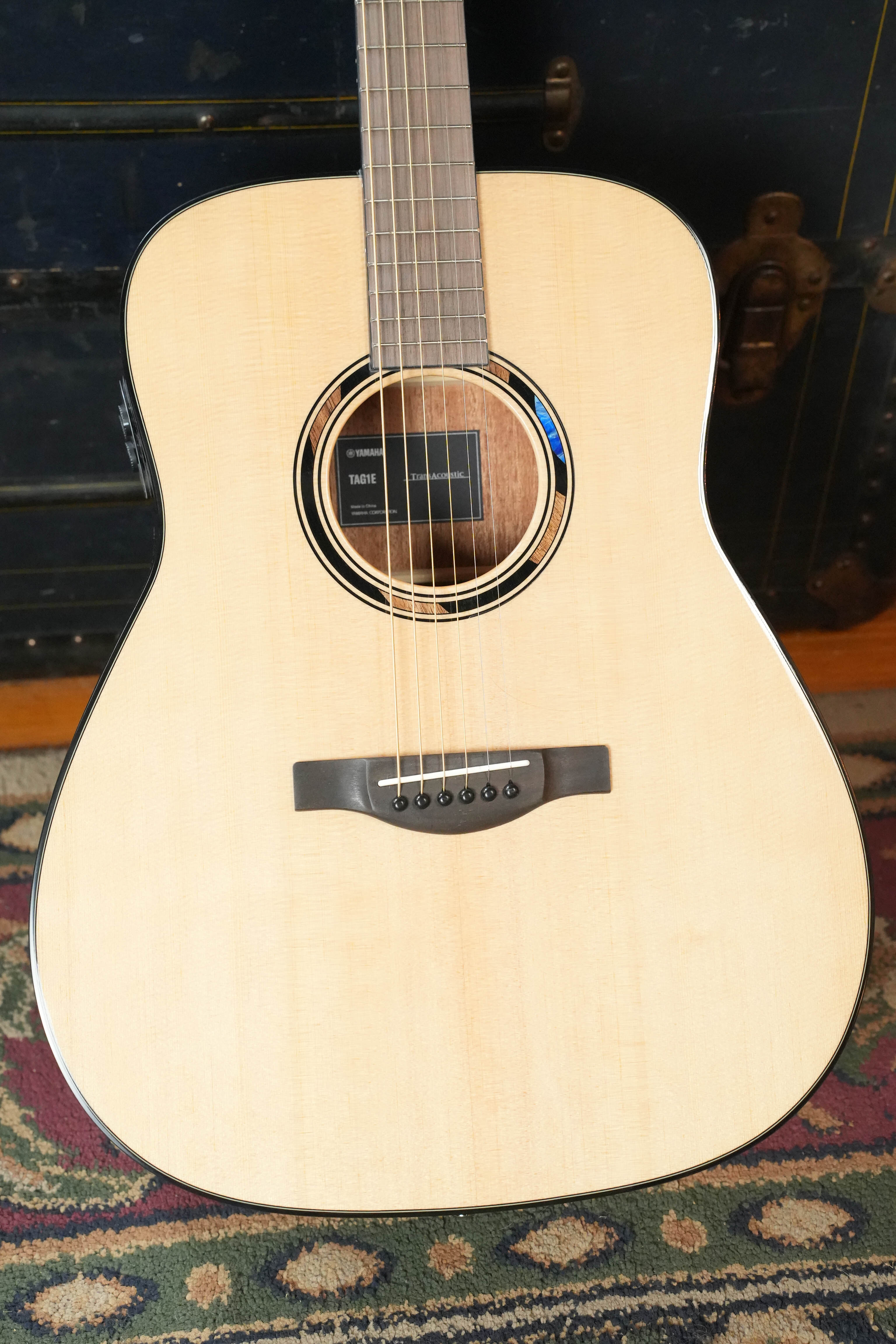 A close-up of the Yamaha TAG1E TransAcoustic Natural Finish Dreadnought Acoustic-Electric Guitar #1116, featuring a light wood body, black bridge, and detailed rosette, rests on a patterned rug—ready to deliver lush acoustic effects.