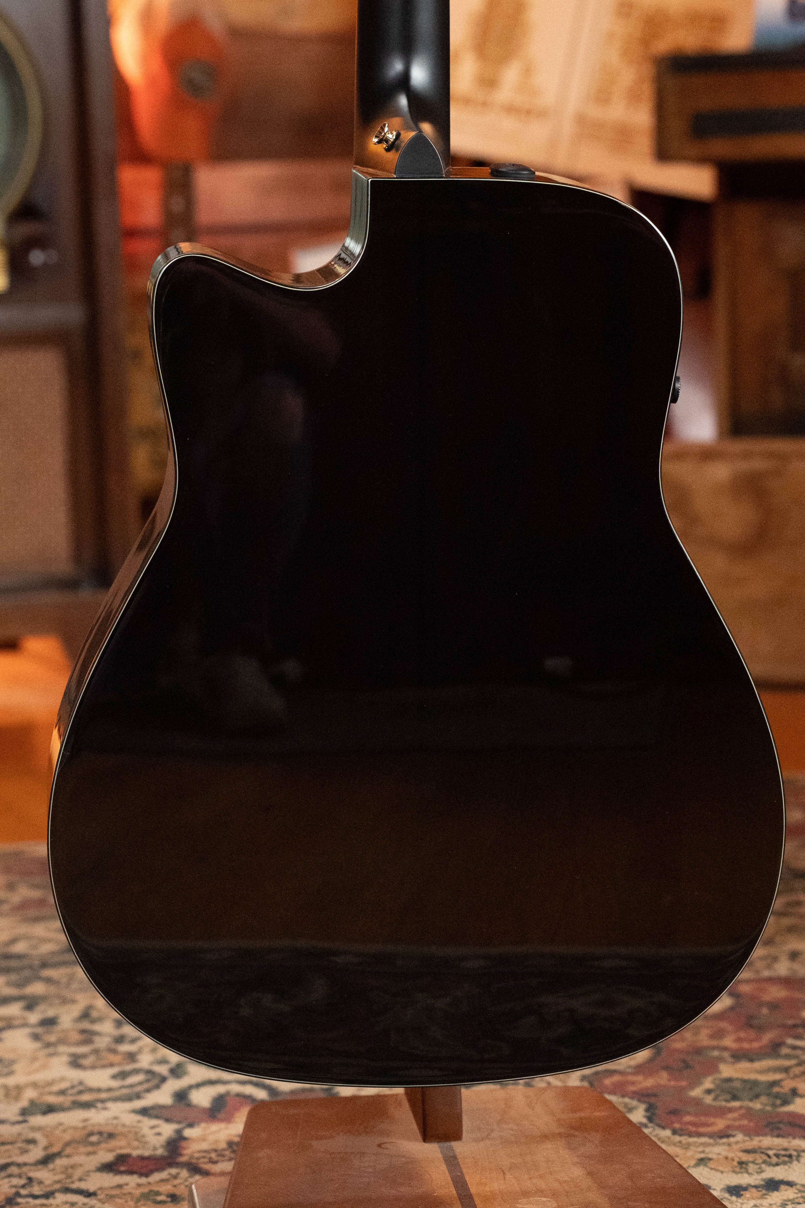 A close-up of the back of a glossy Yamaha TAG3C-NT Sitka/Mahogany Dreadnought Guitar with the newest TransAcoustic System, resting on a wooden stand in a cozy, warmly lit room with a patterned rug.