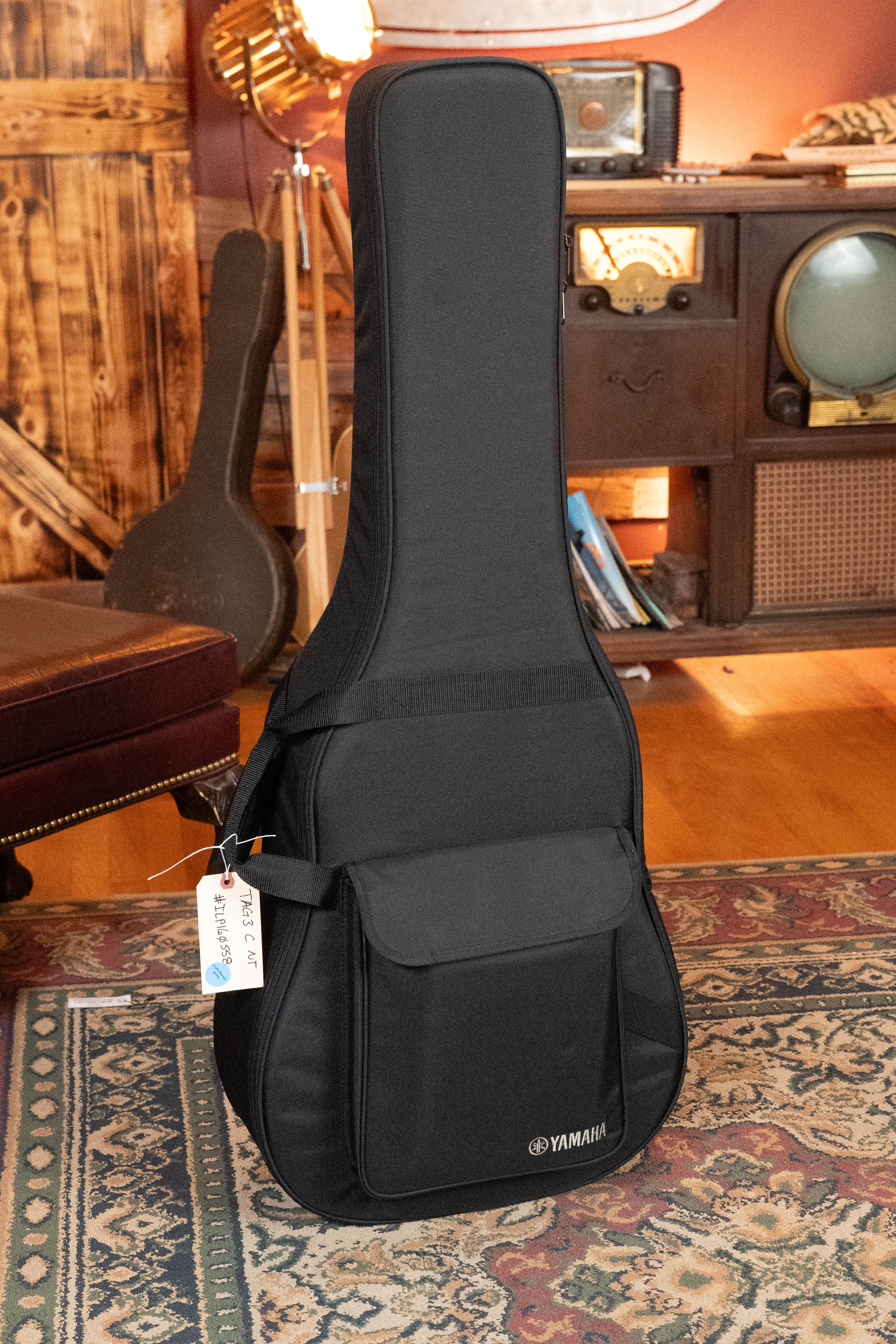 A Yamaha TAG3C-NT Sitka/Mahogany Natural Dreadnought Guitar with the newest TransAcoustic System #0558 rests upright in its black gig bag, featuring a front pocket and white tag, on a patterned rug in a warmly lit vintage room.