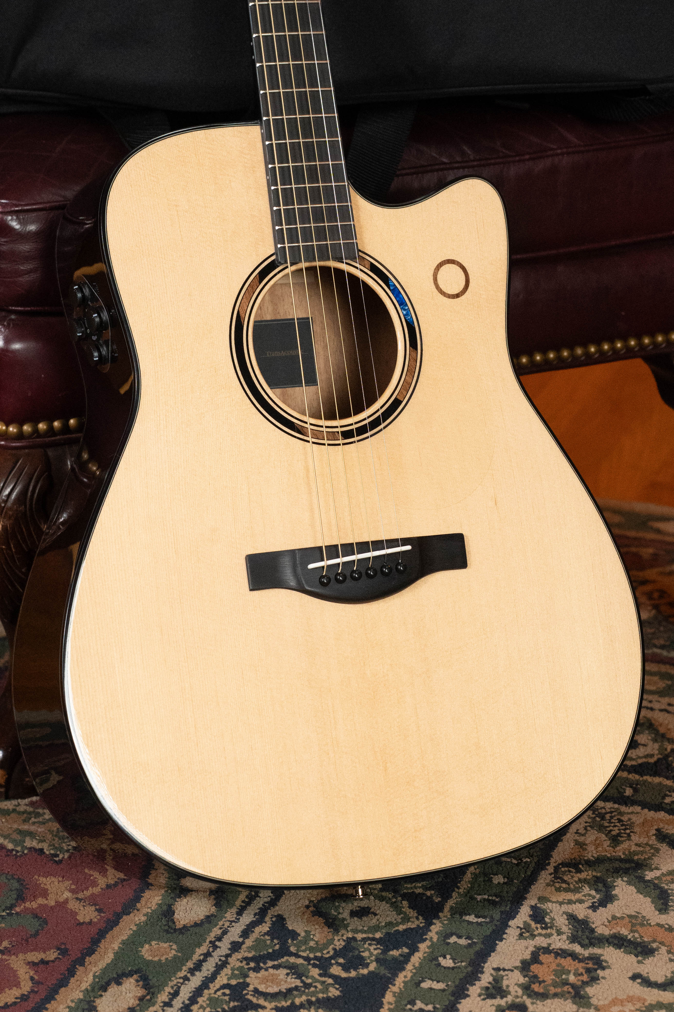 A Yamaha TAG3C-NT Sitka/Mahogany Natural Dreadnought Guitar with the latest TransAcoustic System, featuring a light wood body and black bridge, rests against a leather couch on a patterned rug.